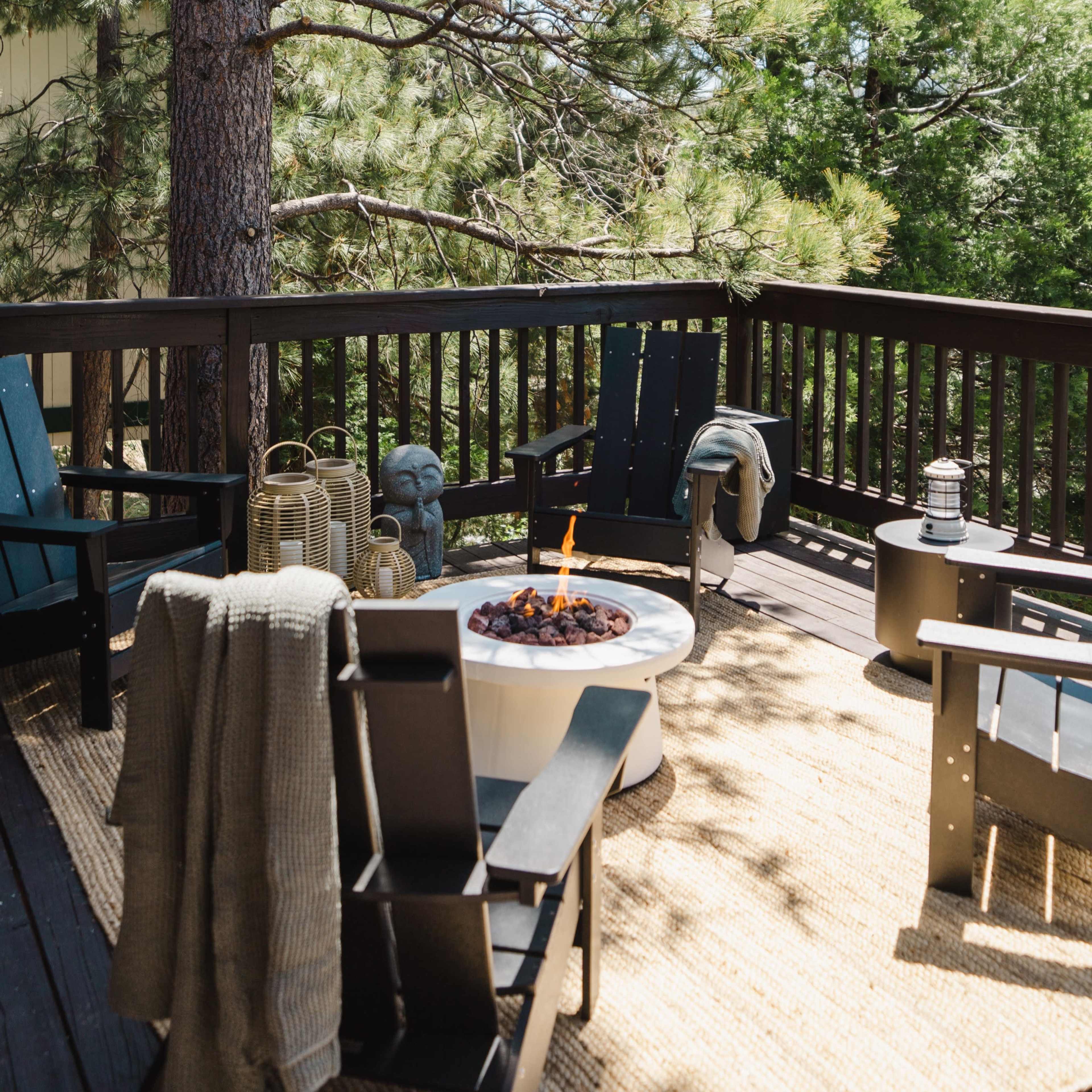 A wooden deck features black outdoor chairs arranged around a fire pit, surrounded by tall trees.
