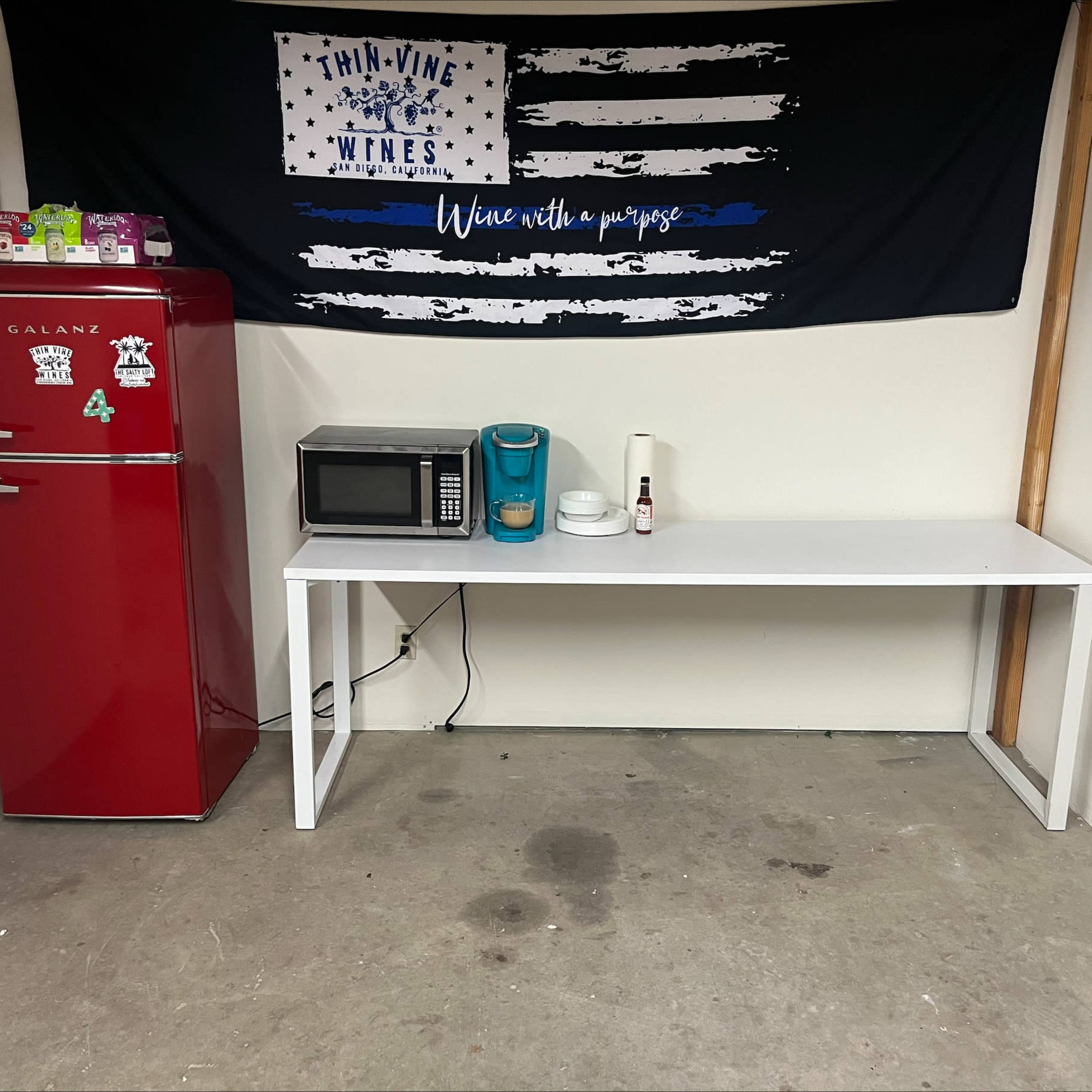 A red refrigerator, a microwave, and a coffee maker on a white table, with a black banner featuring a blue line American flag hanging above.