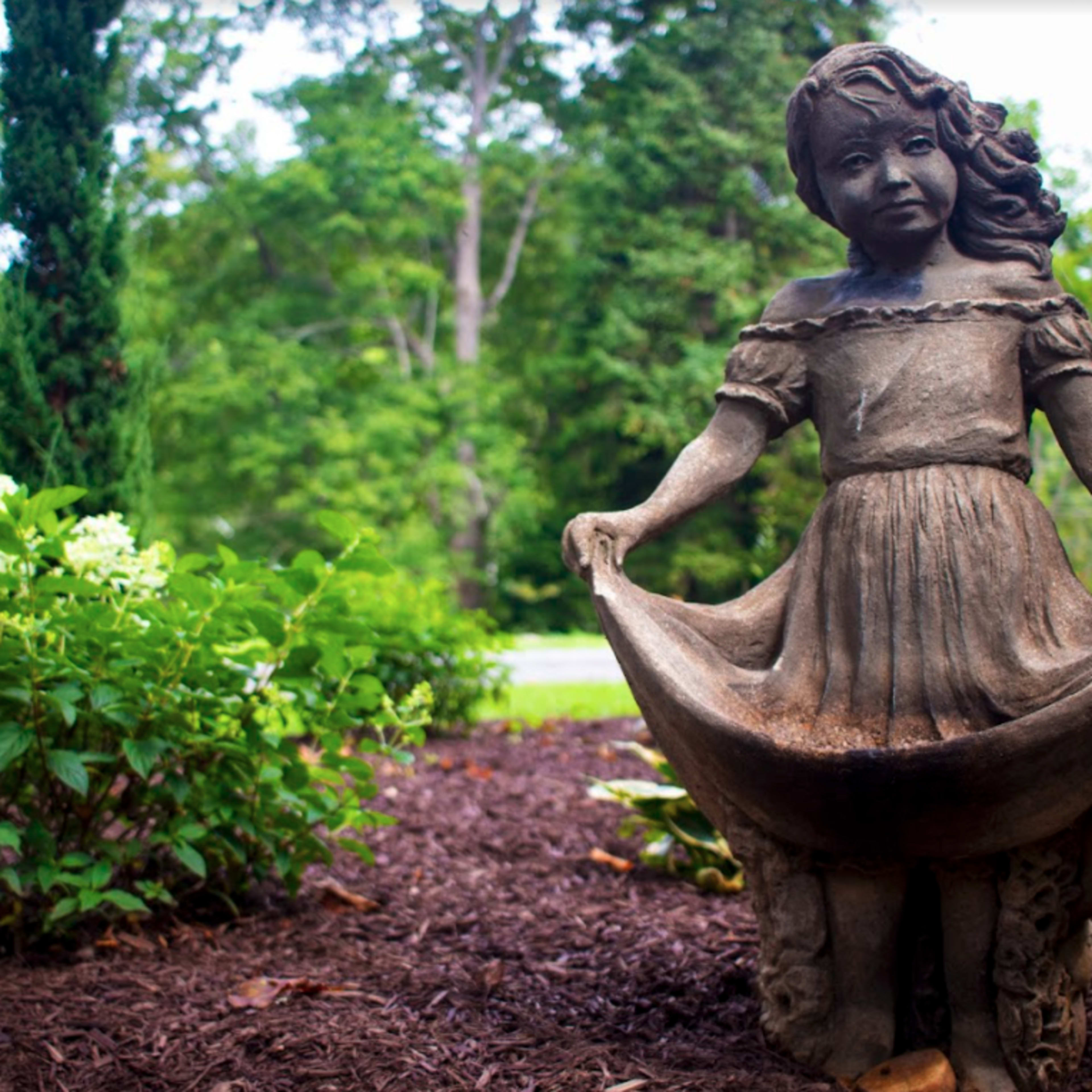 A concrete statue of a girl holding her skirt stands among green foliage and mulch in a garden.