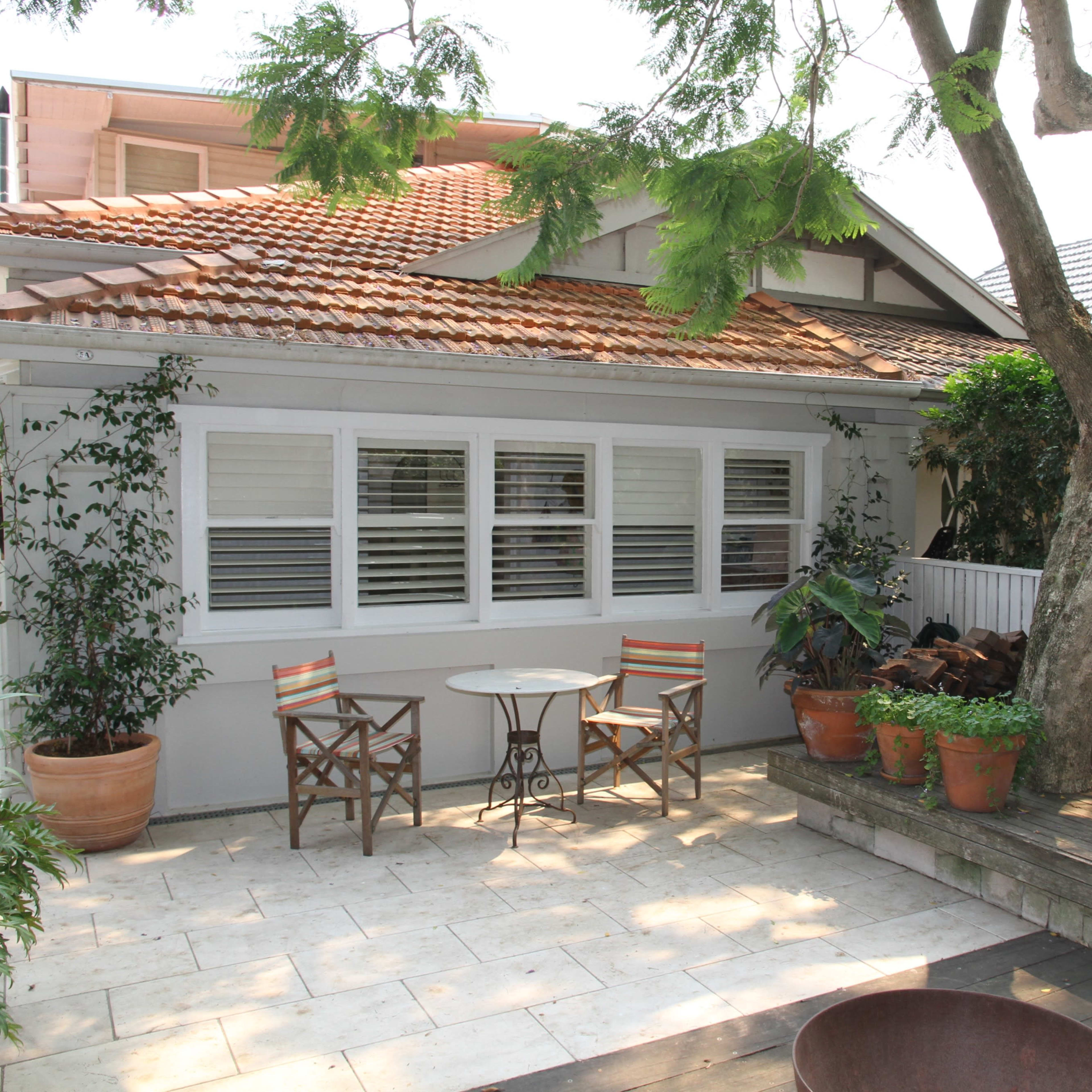 A patio area features two chairs and a small table beside a house with a tiled roof, surrounded by potted plants and a large tree.
