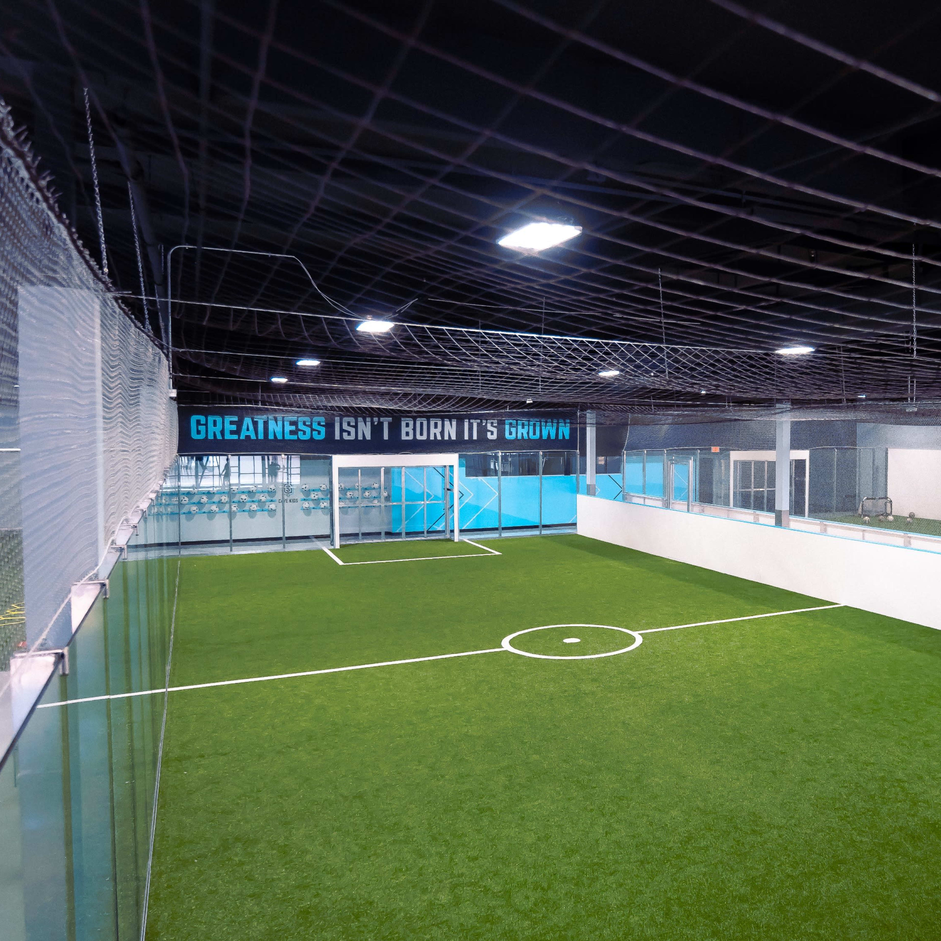An indoor soccer facility features a well-maintained artificial turf field surrounded by walls and nets.