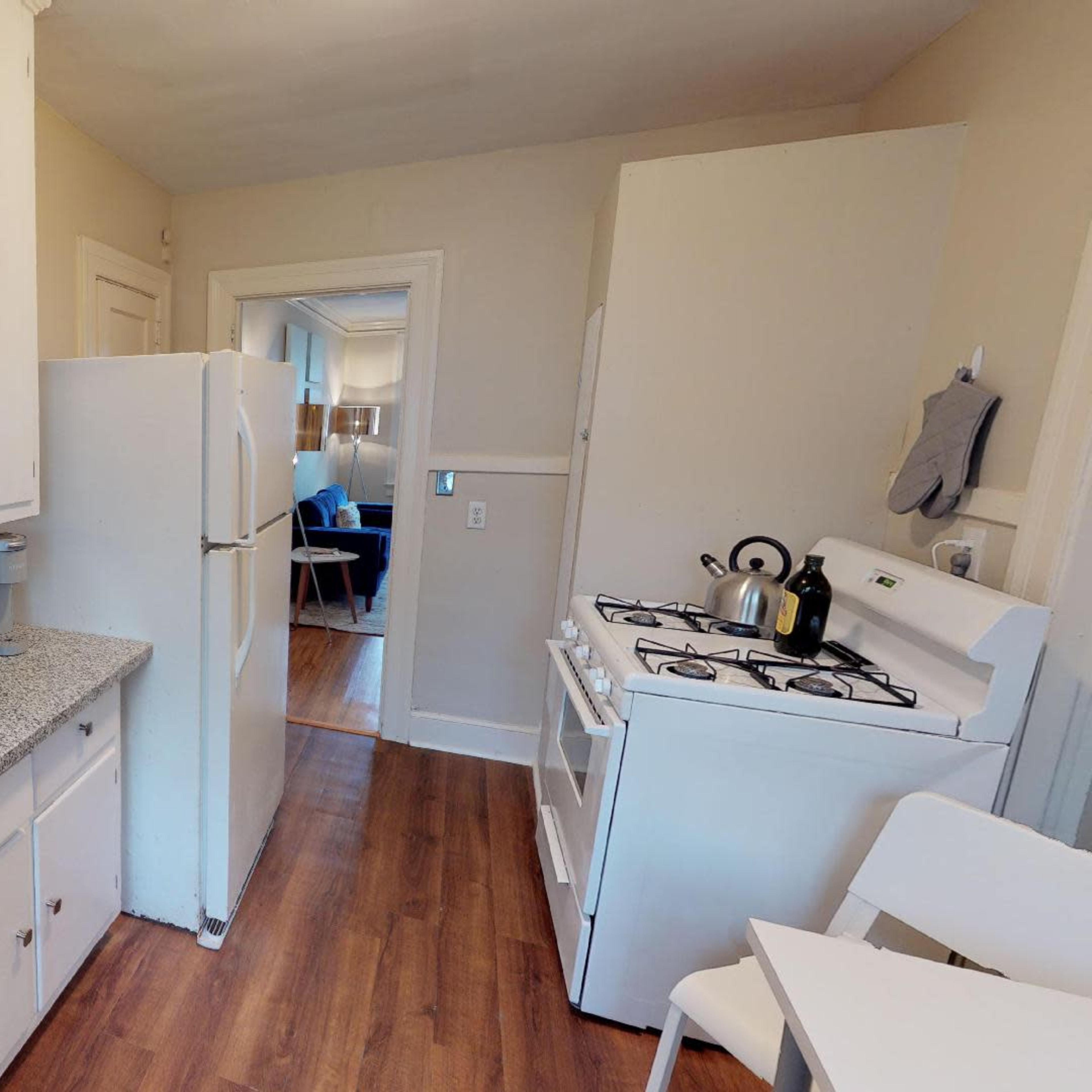 A compact kitchen featuring white cabinets, a sink, a stove, and a refrigerator, with wooden flooring and a glimpse of a living area in the background.
