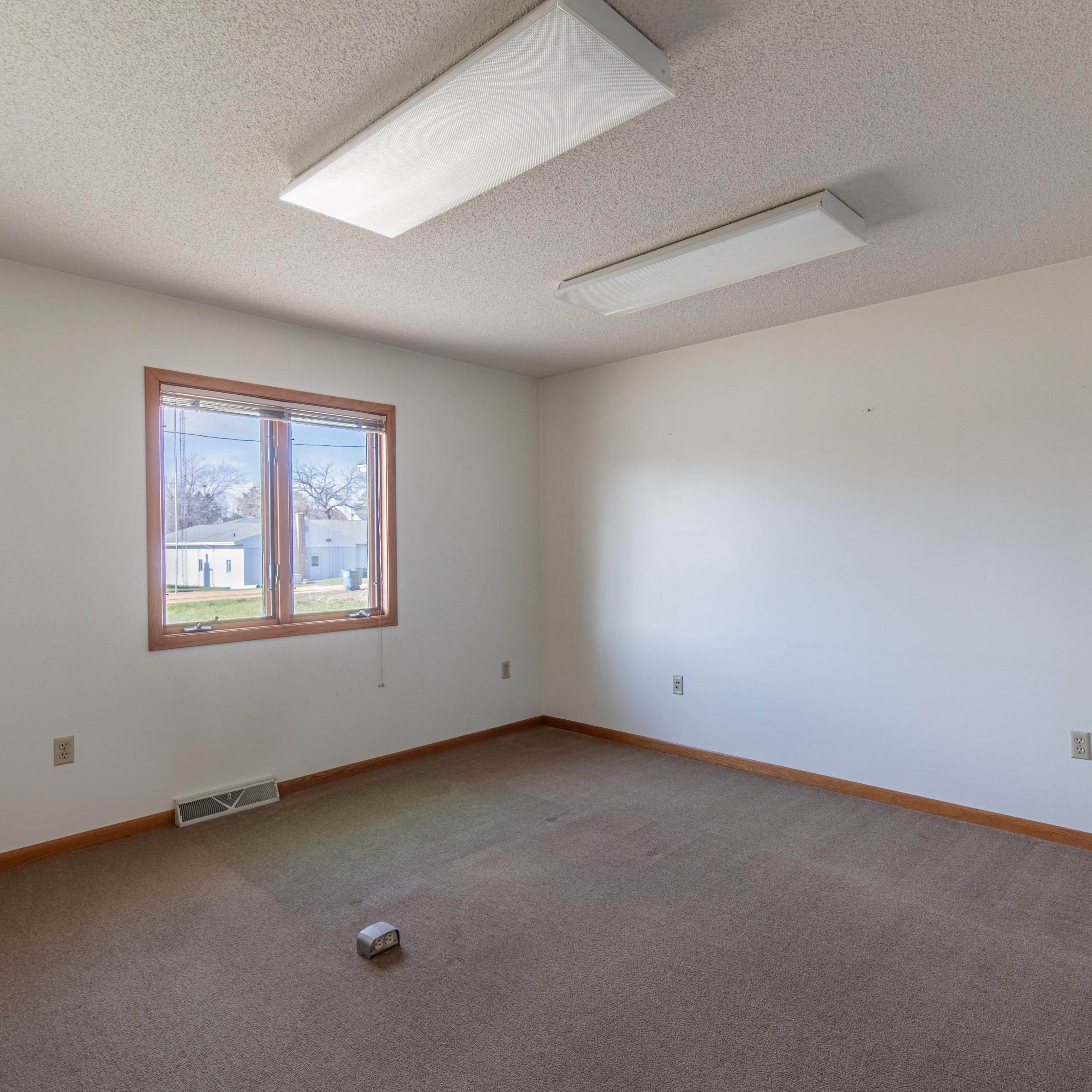 An empty room with beige carpet, white walls, and a single window allows natural light to enter.