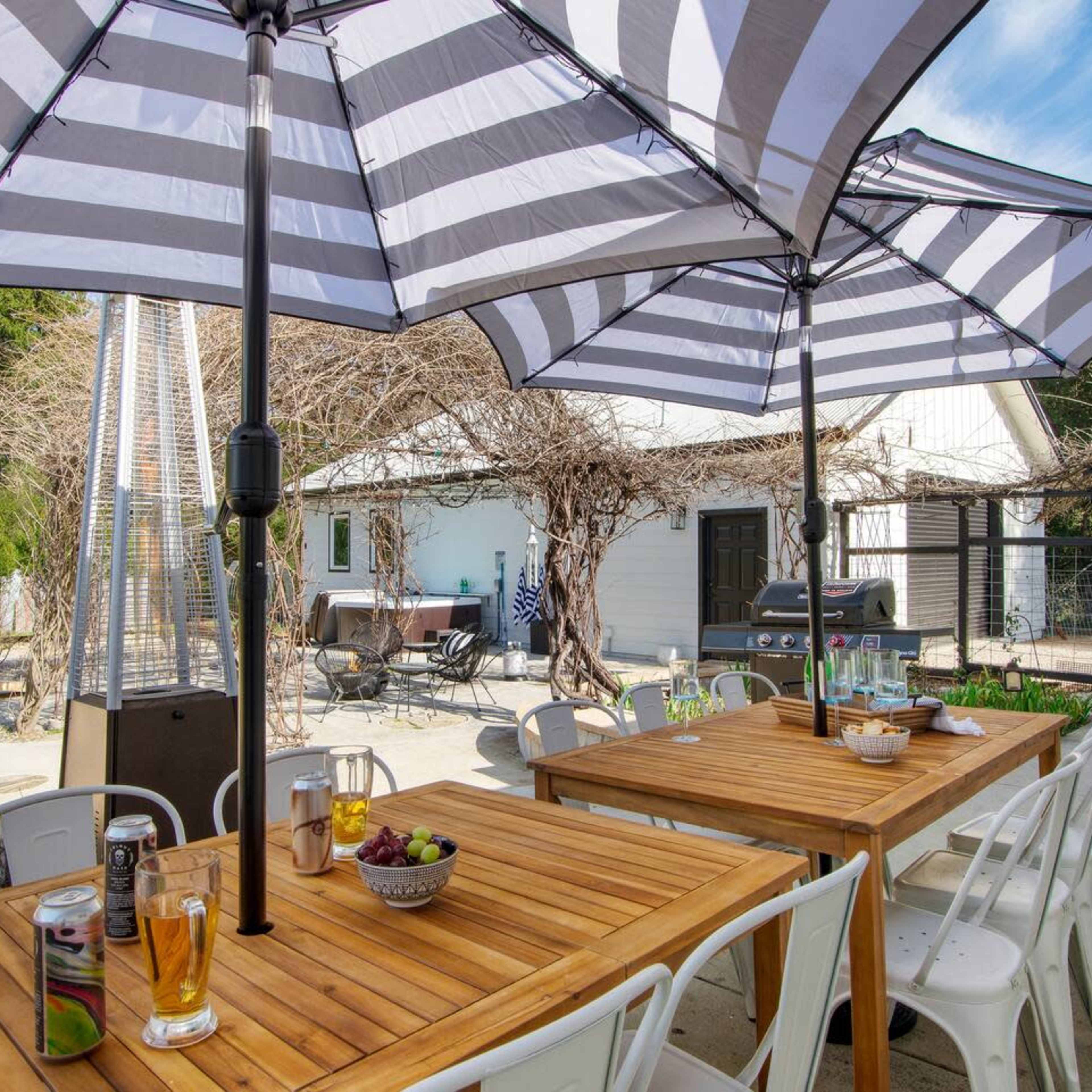 The scene features a spacious outdoor dining area with wooden tables, striped umbrellas, and a backdrop of a house surrounded by greenery.