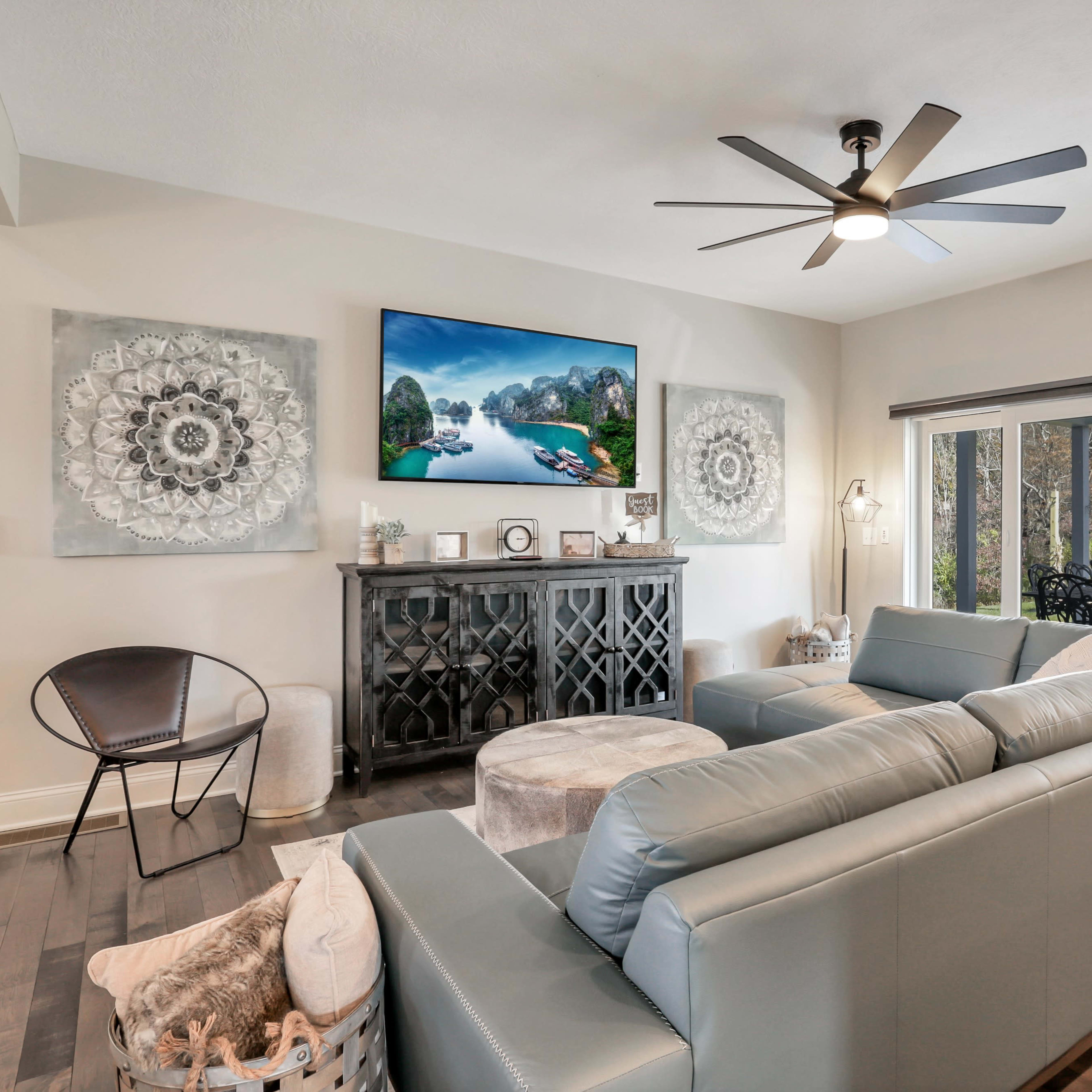 A modern living room features a sectional sofa, a circular ottoman, a sleek media console, and large windows that open to a patio.