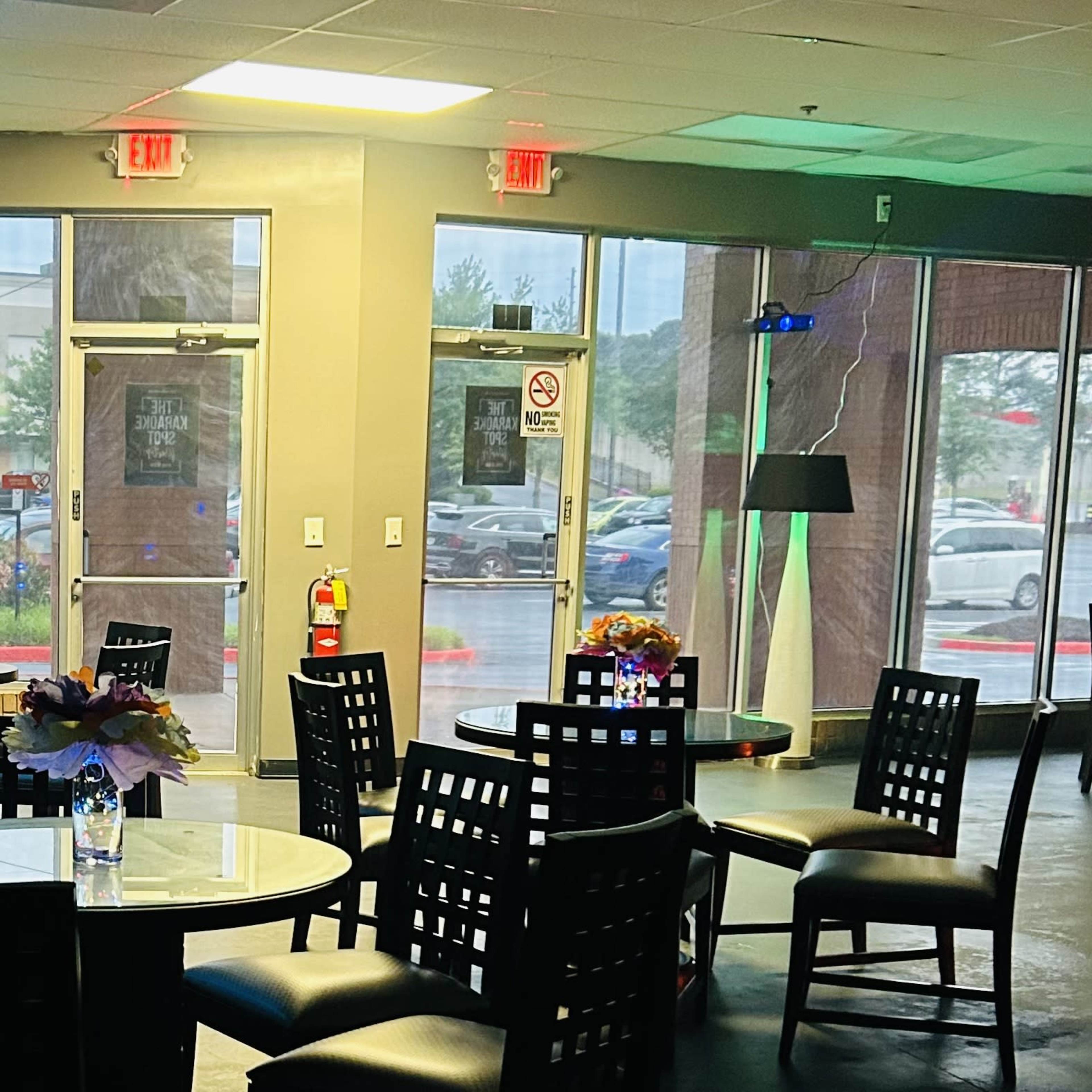 A dining area with several tables and chairs arranged around small floral centerpieces, illuminated by natural light from large windows.