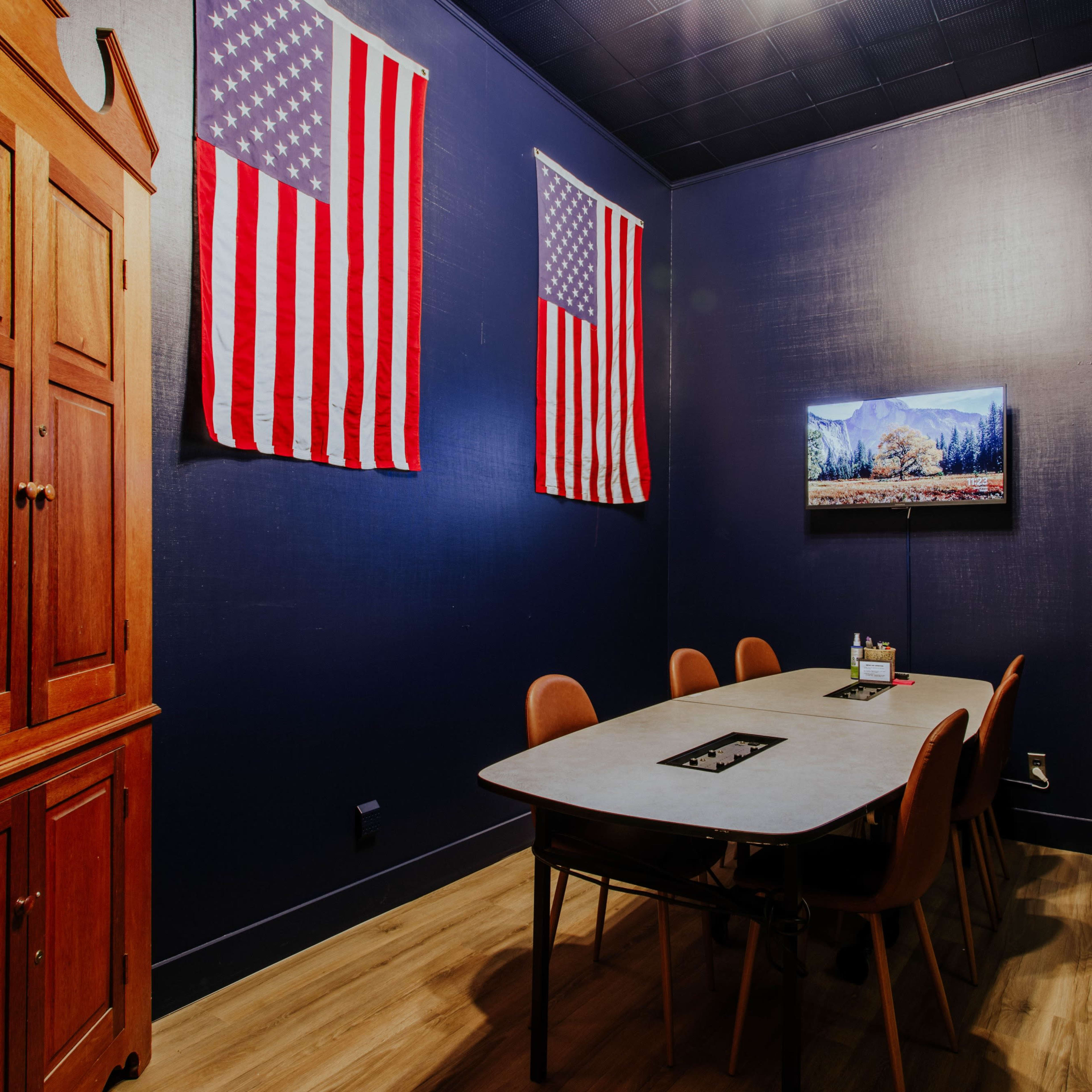 A conference room with a wooden cabinet, a rectangular table surrounded by six chairs, two American flags on the walls, and a television mounted on one wall.