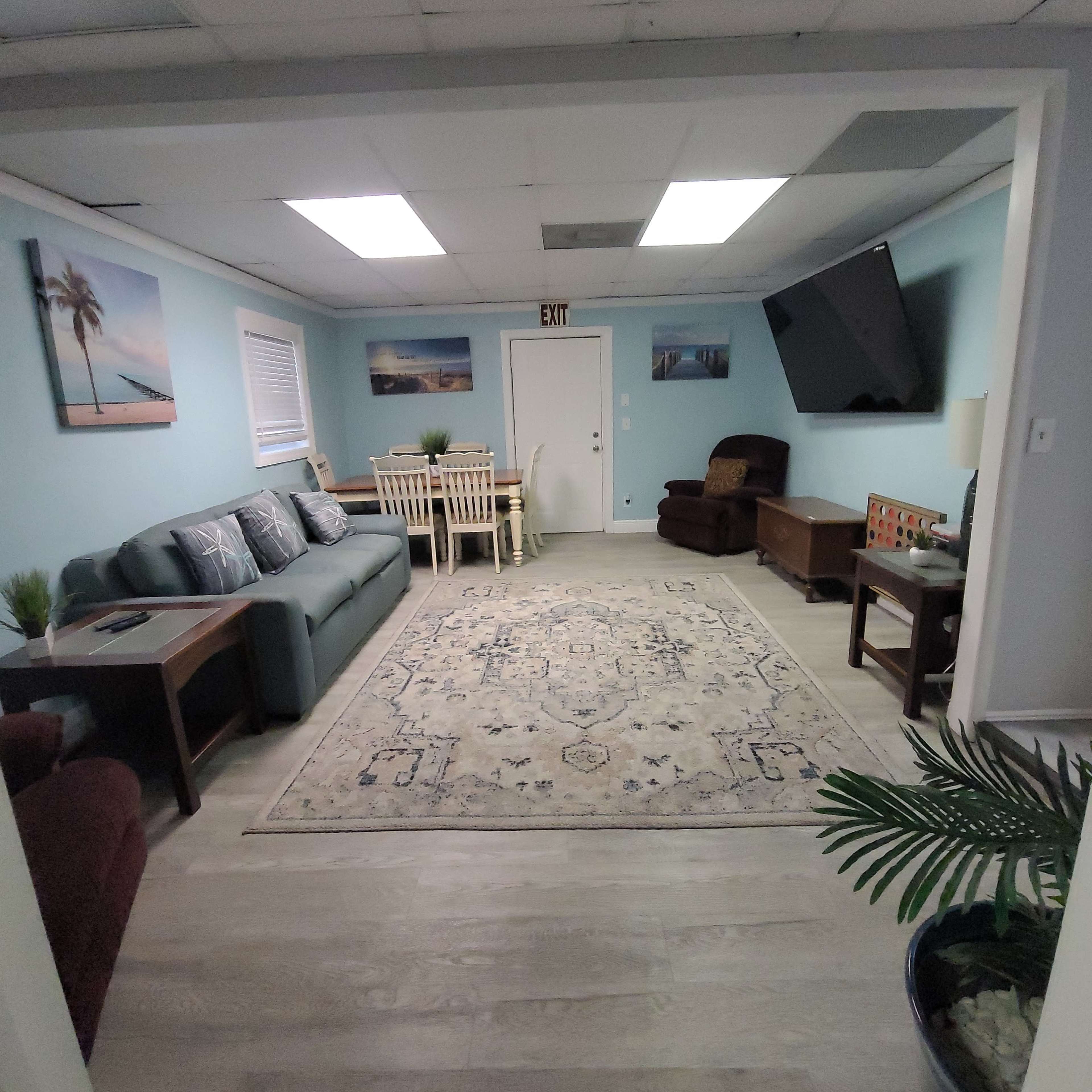 A living room area with a blue walls, a large area rug, a sofa set, a dining table, and a wall-mounted television.
