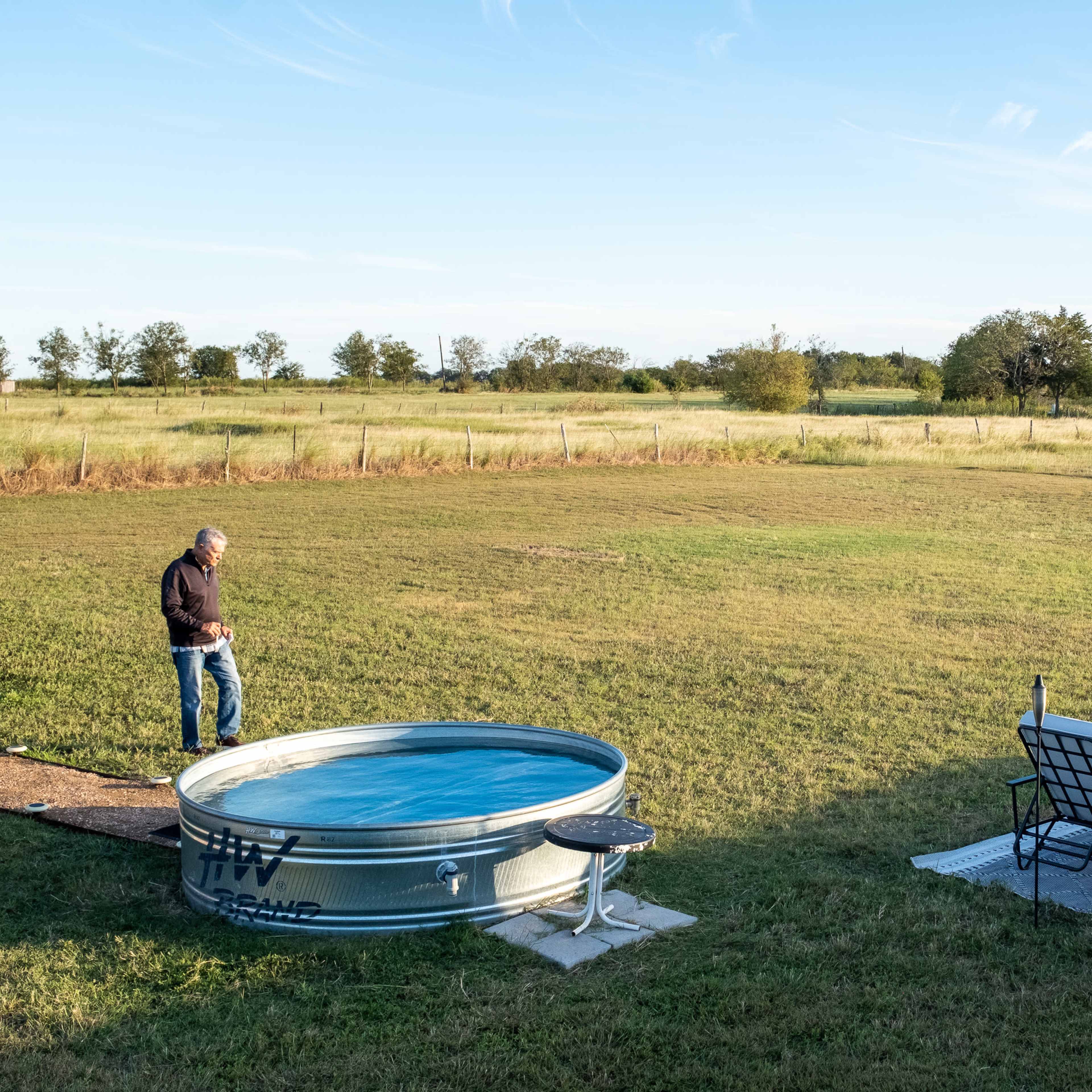 A man stands near a metal stock tank pool, while a woman sits on a patio adjacent to two chairs in a grassy field.