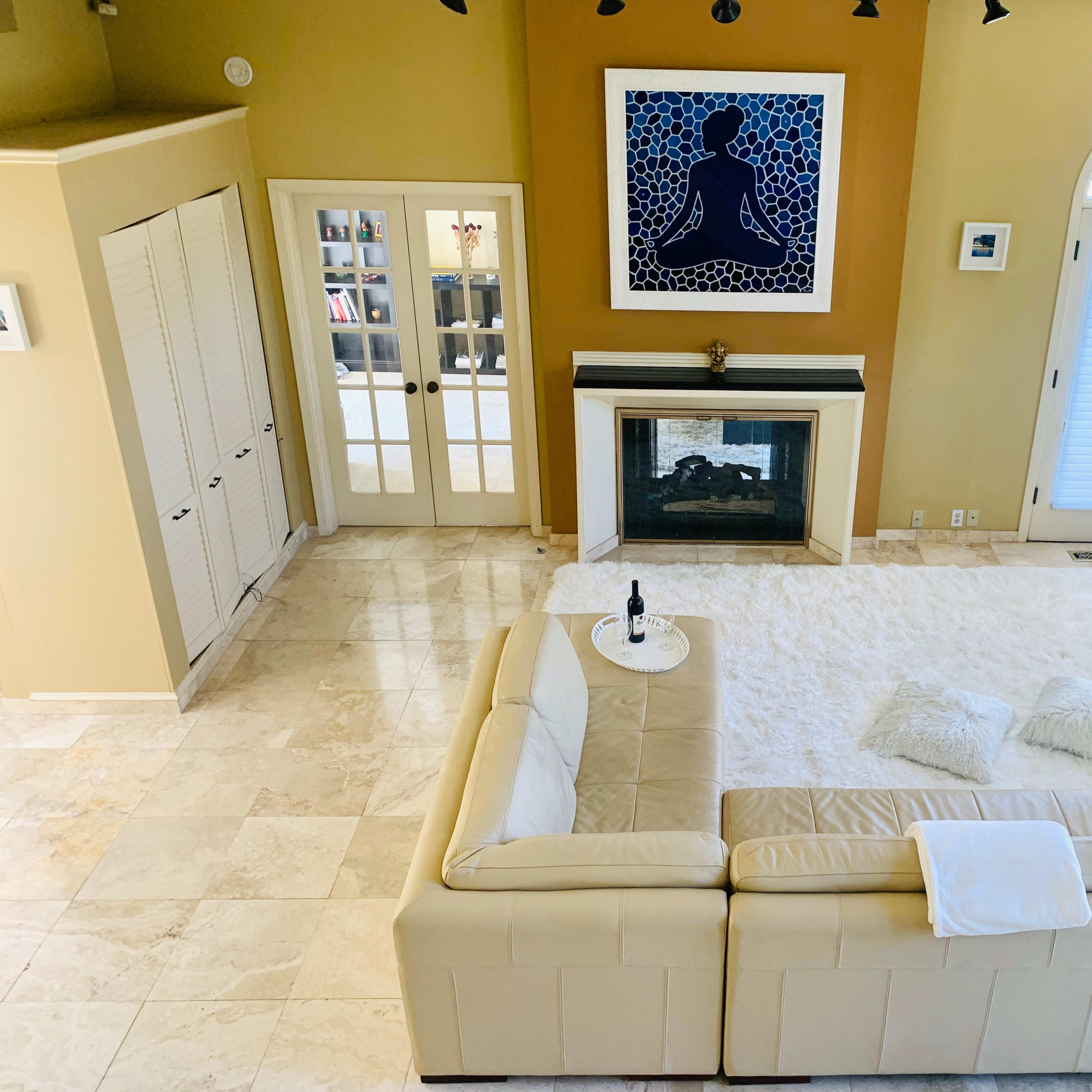 The room features a large beige sectional sofa, a white area rug, a fireplace, and a framed artwork on the wall, with double doors leading to another room.