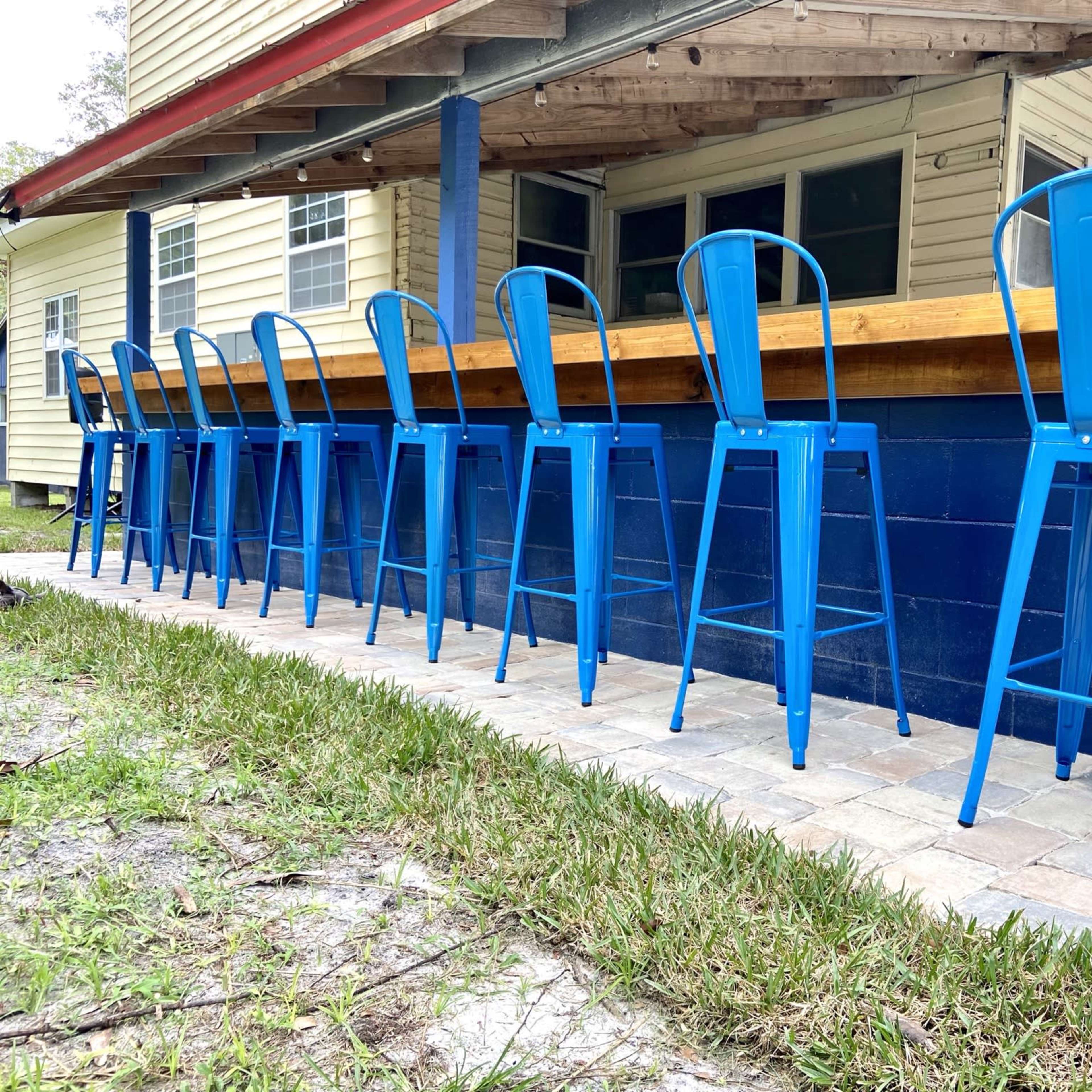 A row of blue metal bar stools is aligned along a wooden counter outside a building with yellow siding.