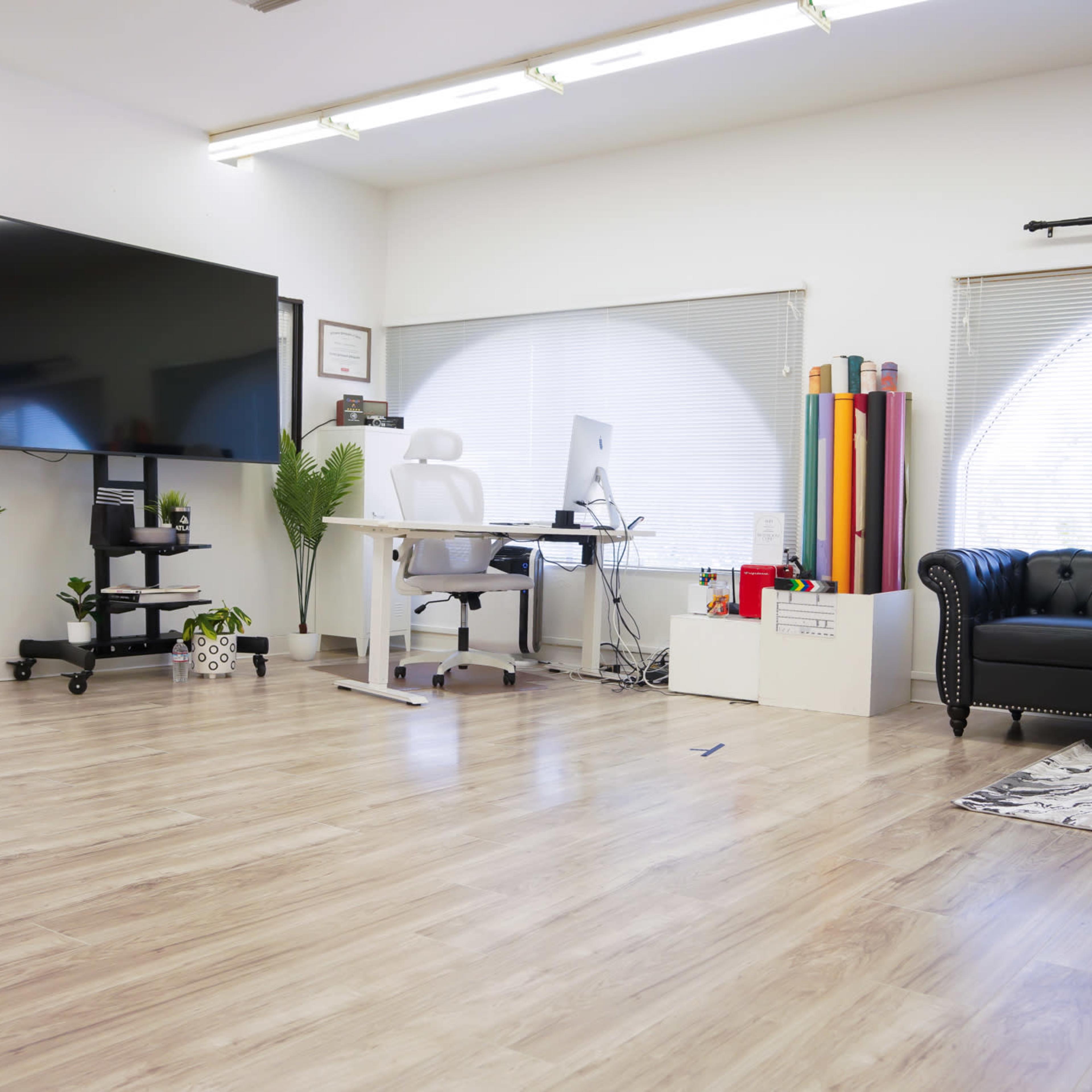 A modern office space featuring a large TV mounted on the wall, a white desk with a computer, a black couch, and various indoor plants.