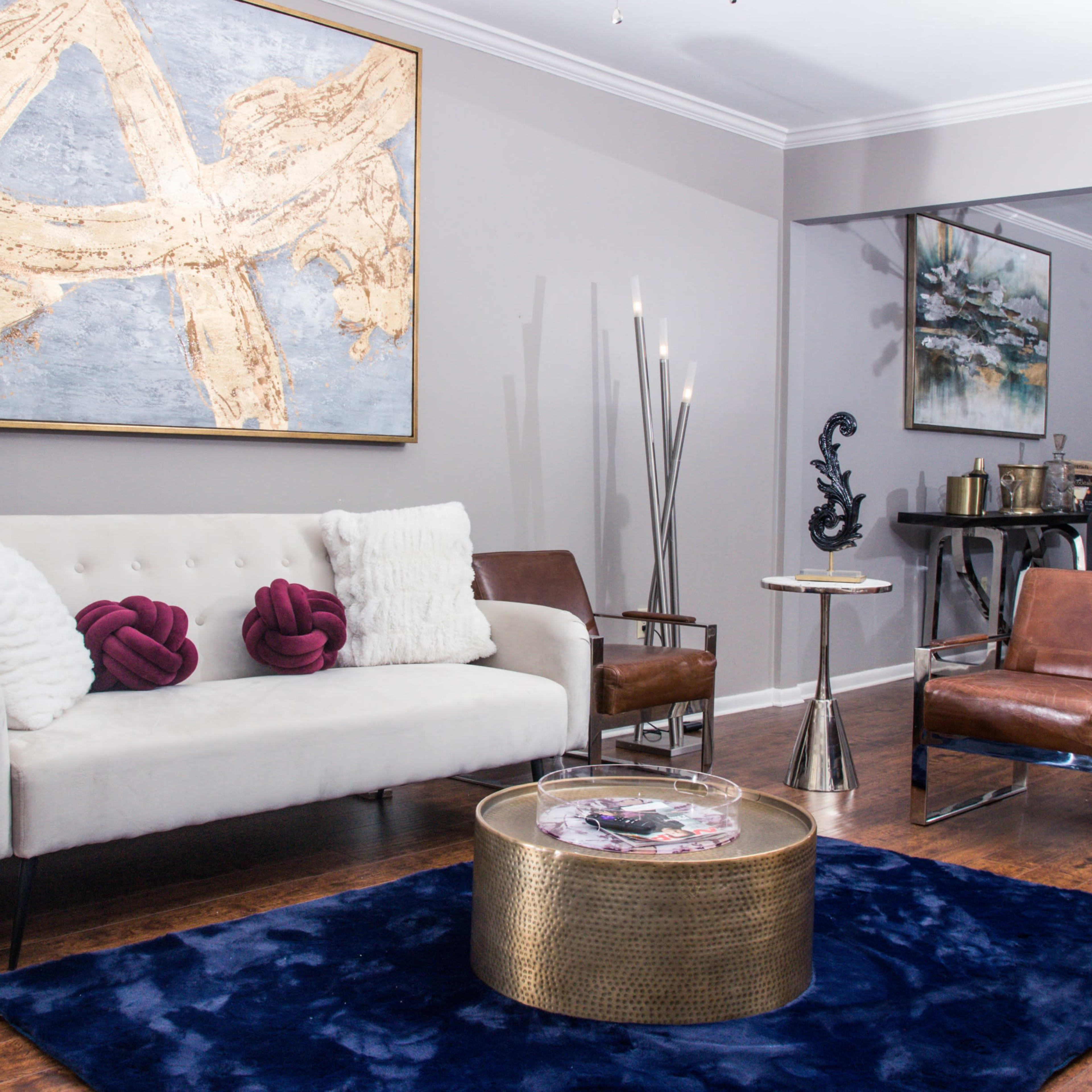 A modern living room featuring a white sofa, brown accent chairs, a dark blue rug, and contemporary artwork on the walls.