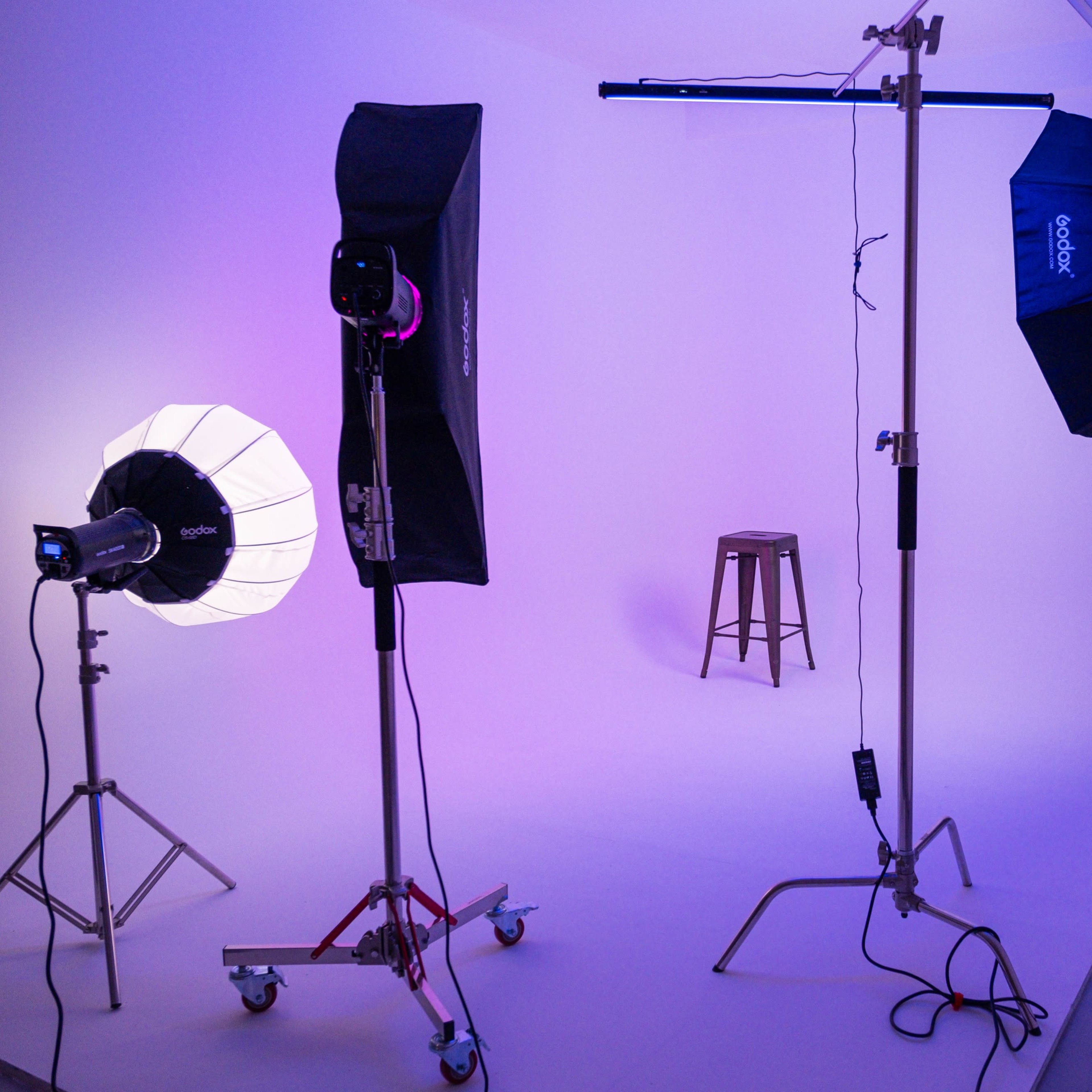 A photography studio setup with multiple light sources, including a softbox and an umbrella light, and a wooden stool in the center against a white backdrop.