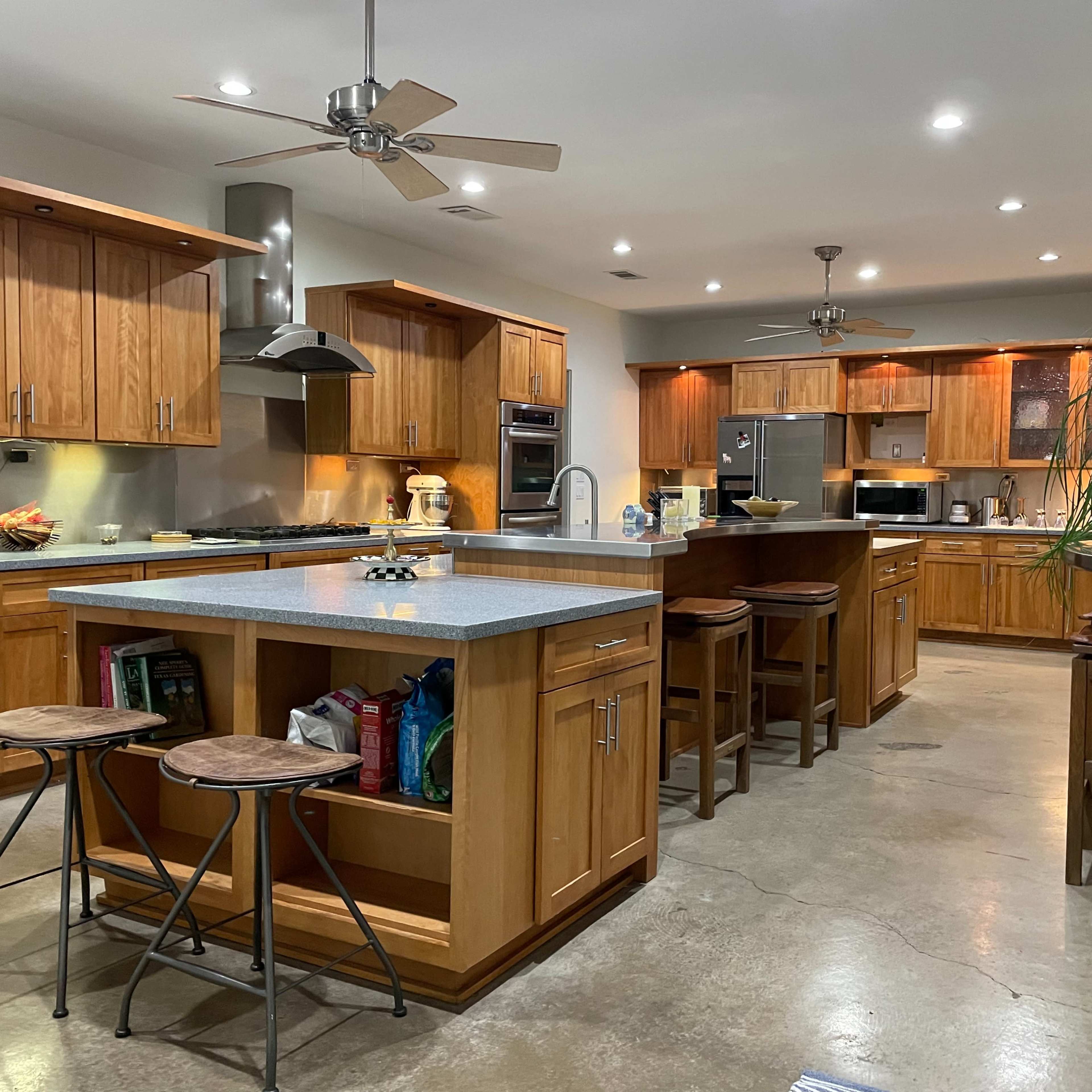 A modern kitchen featuring wooden cabinets, stainless steel appliances, a large island with bar stools, and a concrete floor.