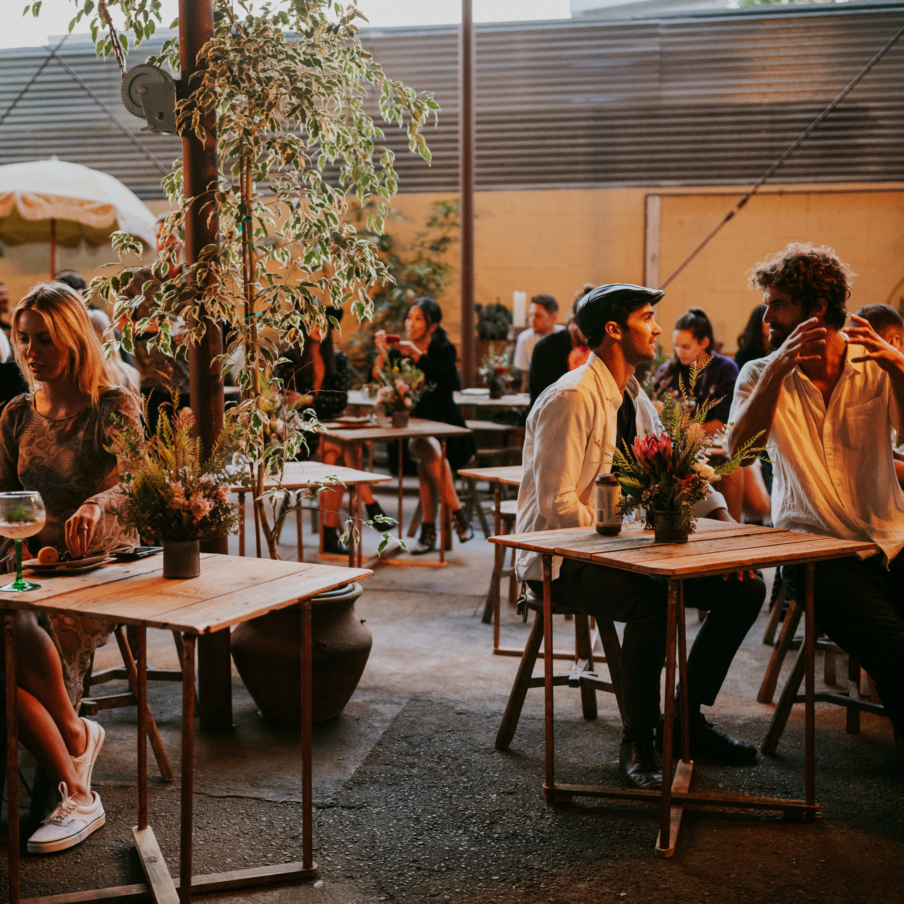 A lively gathering in an outdoor café with people seated at wooden tables surrounded by plants.