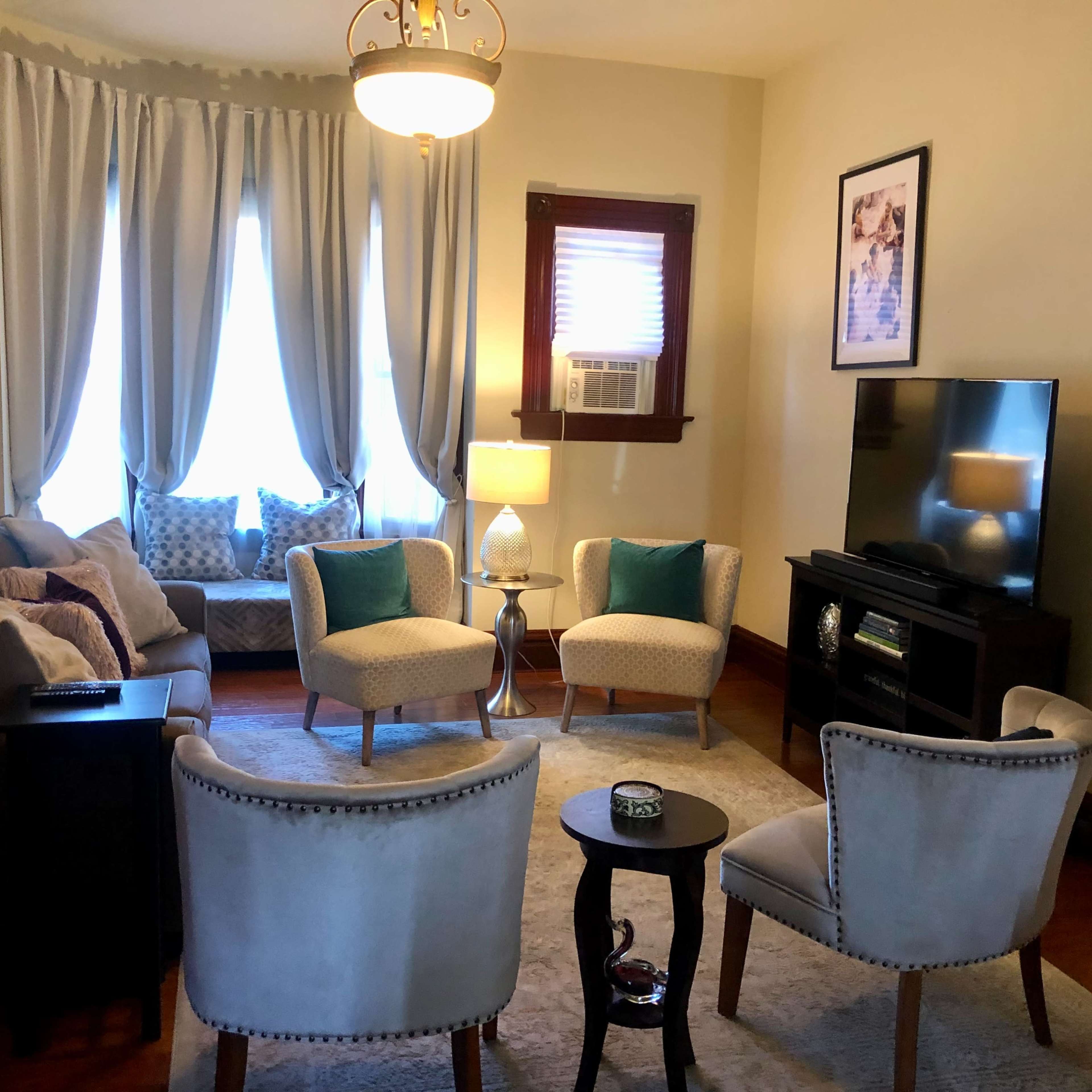 A cozy living room with two armchairs and a sofa arranged around a small table, a television against the wall, and large windows dressed with light curtains.
