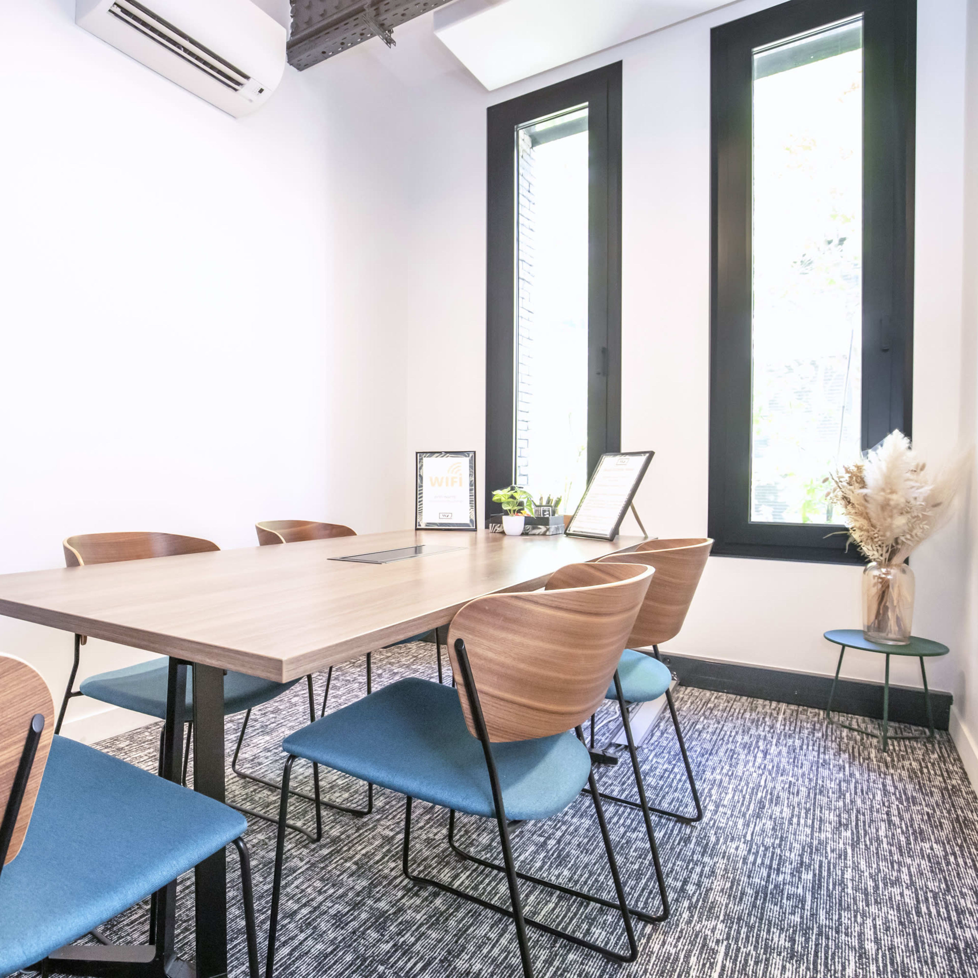 A modern conference room features a wooden table surrounded by six chairs, with large windows allowing natural light in.