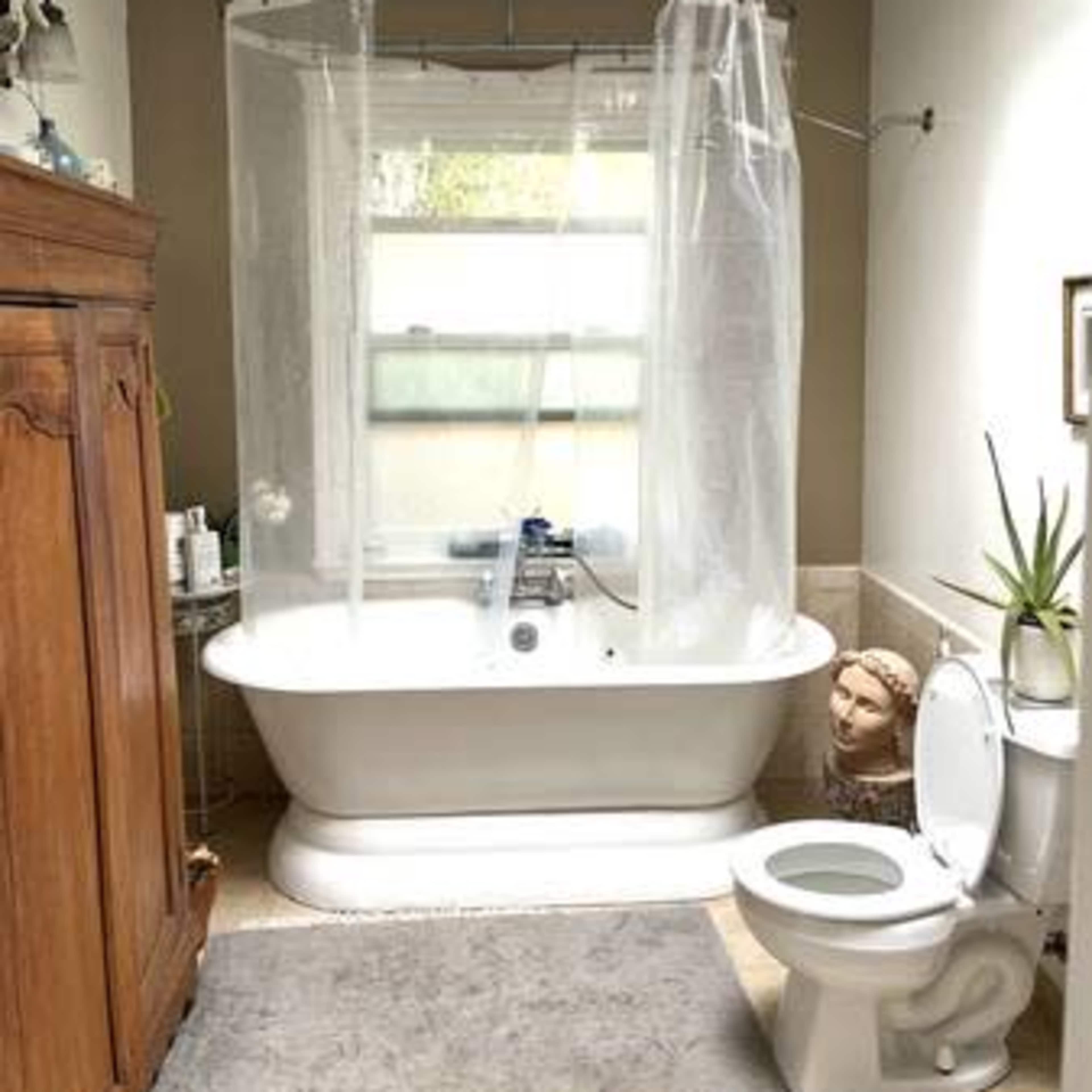 A bathroom features a freestanding bathtub with a shower curtain, a toilet, and a wooden armoire against the wall.