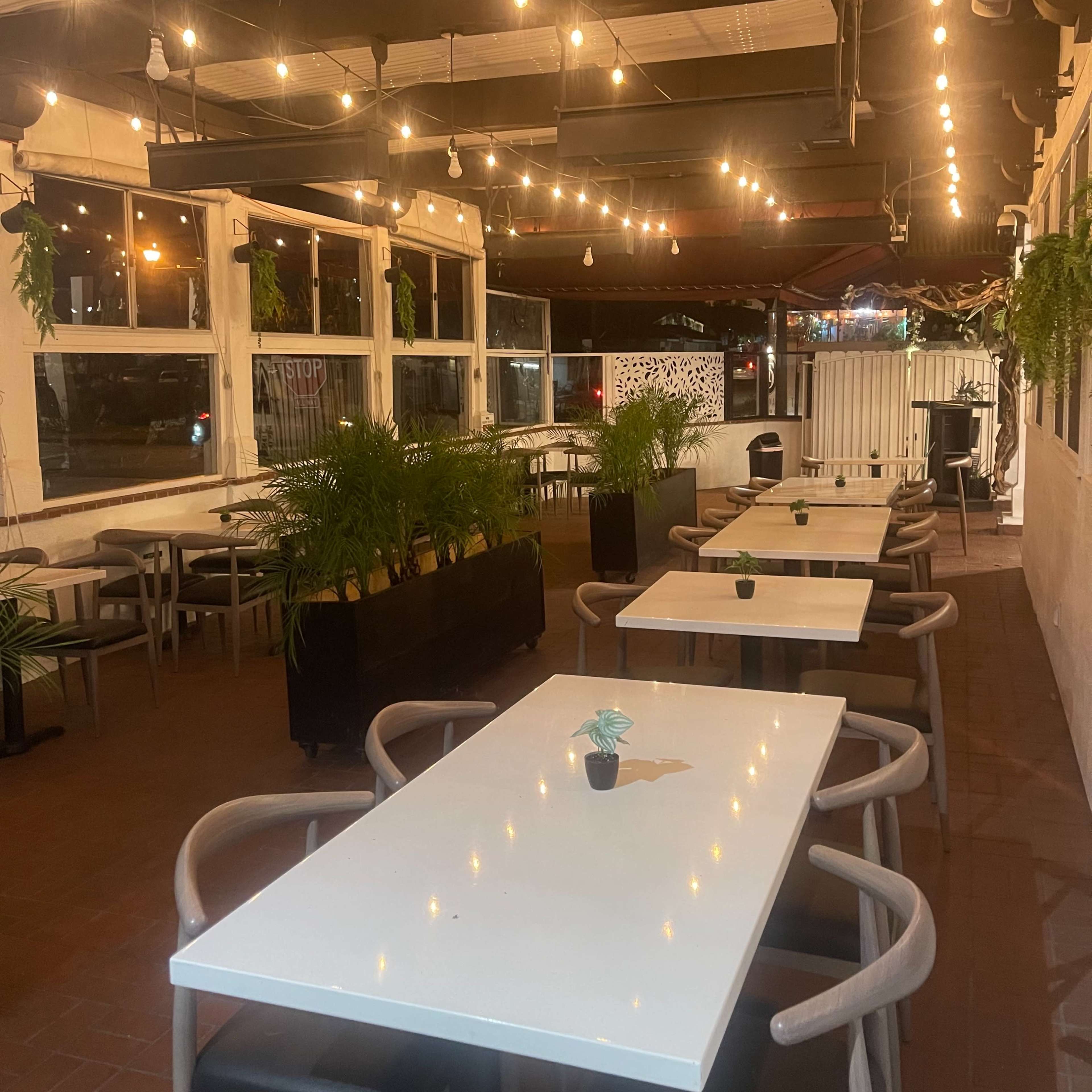 An outdoor restaurant space with several white tables, string lights overhead, and potted plants along the walls.