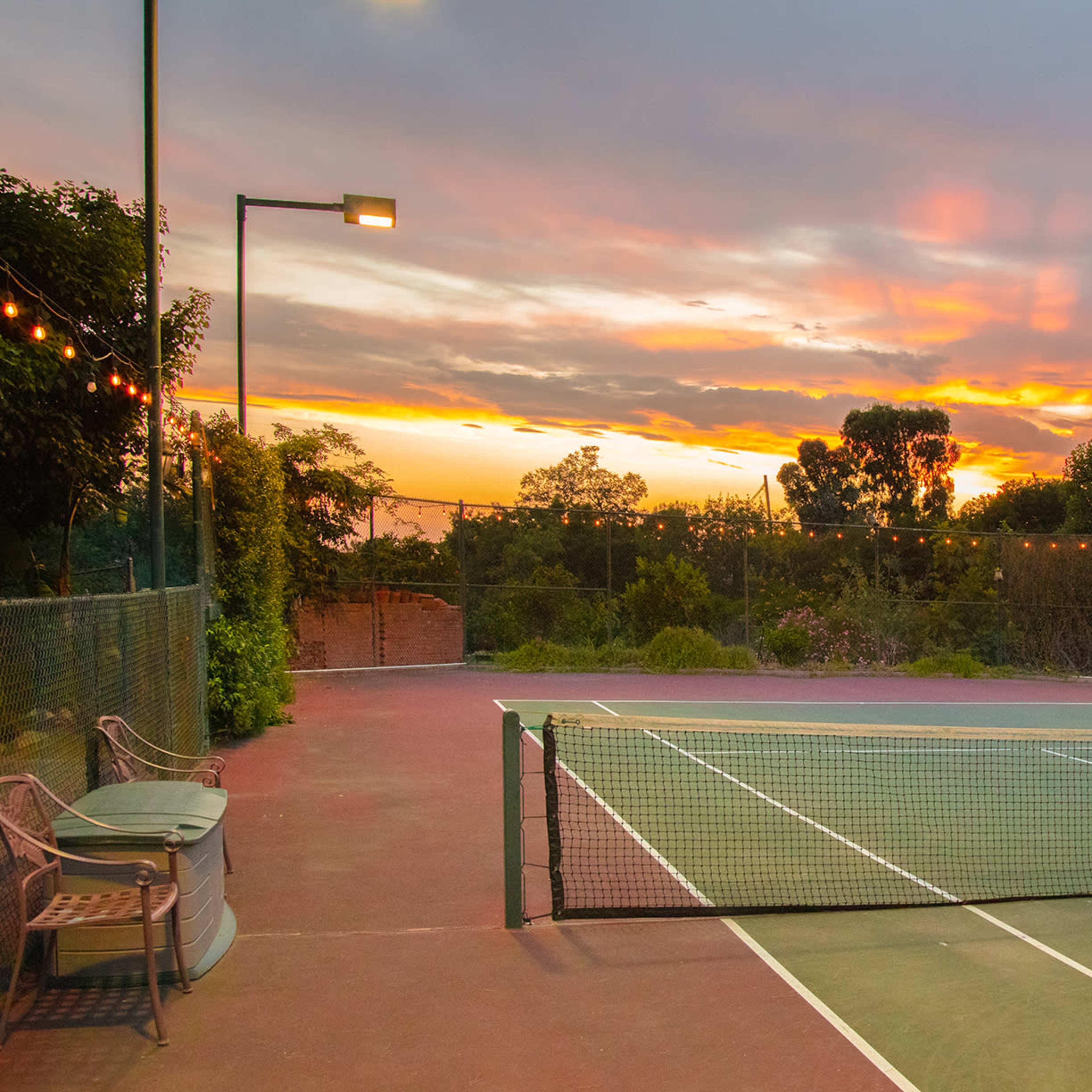 A tennis court is shown at sunset, with vibrant colors in the sky and palm trees in the background.