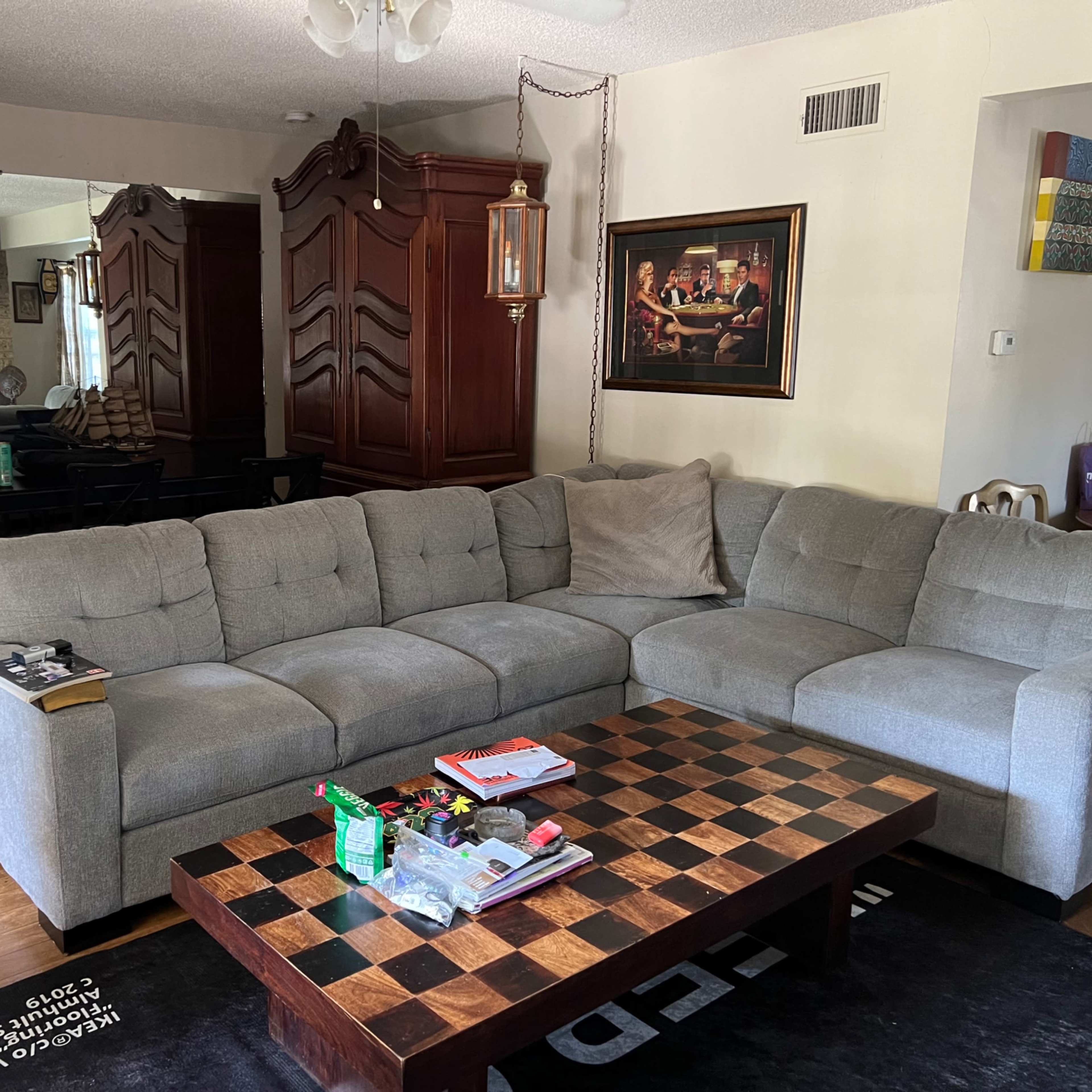 A living room featuring a gray sectional sofa, a chessboard coffee table, and a dining area with a wooden table in the background.