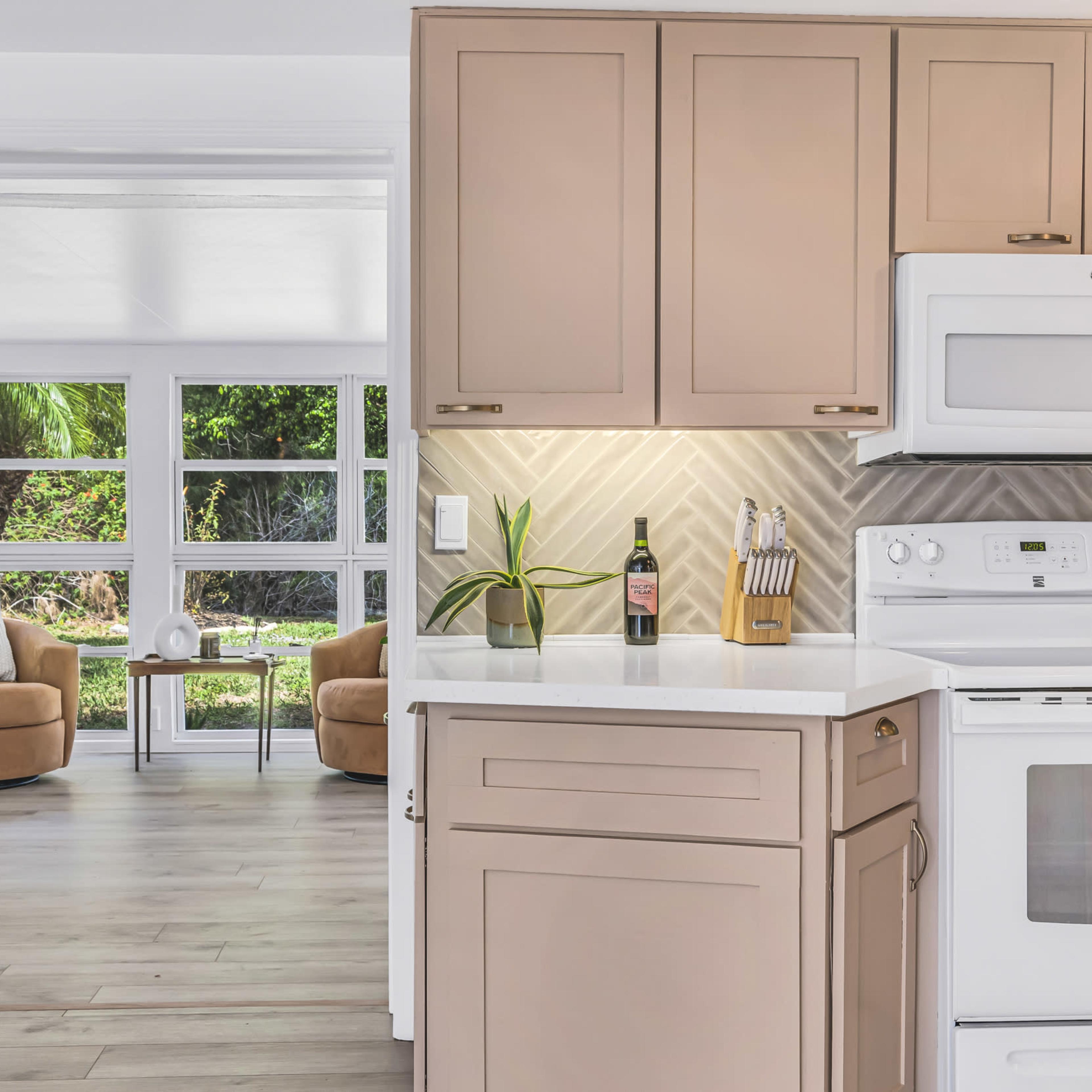 A modern kitchen with light cabinetry and a white stove, overlooking a bright living area with two armchairs and large windows.