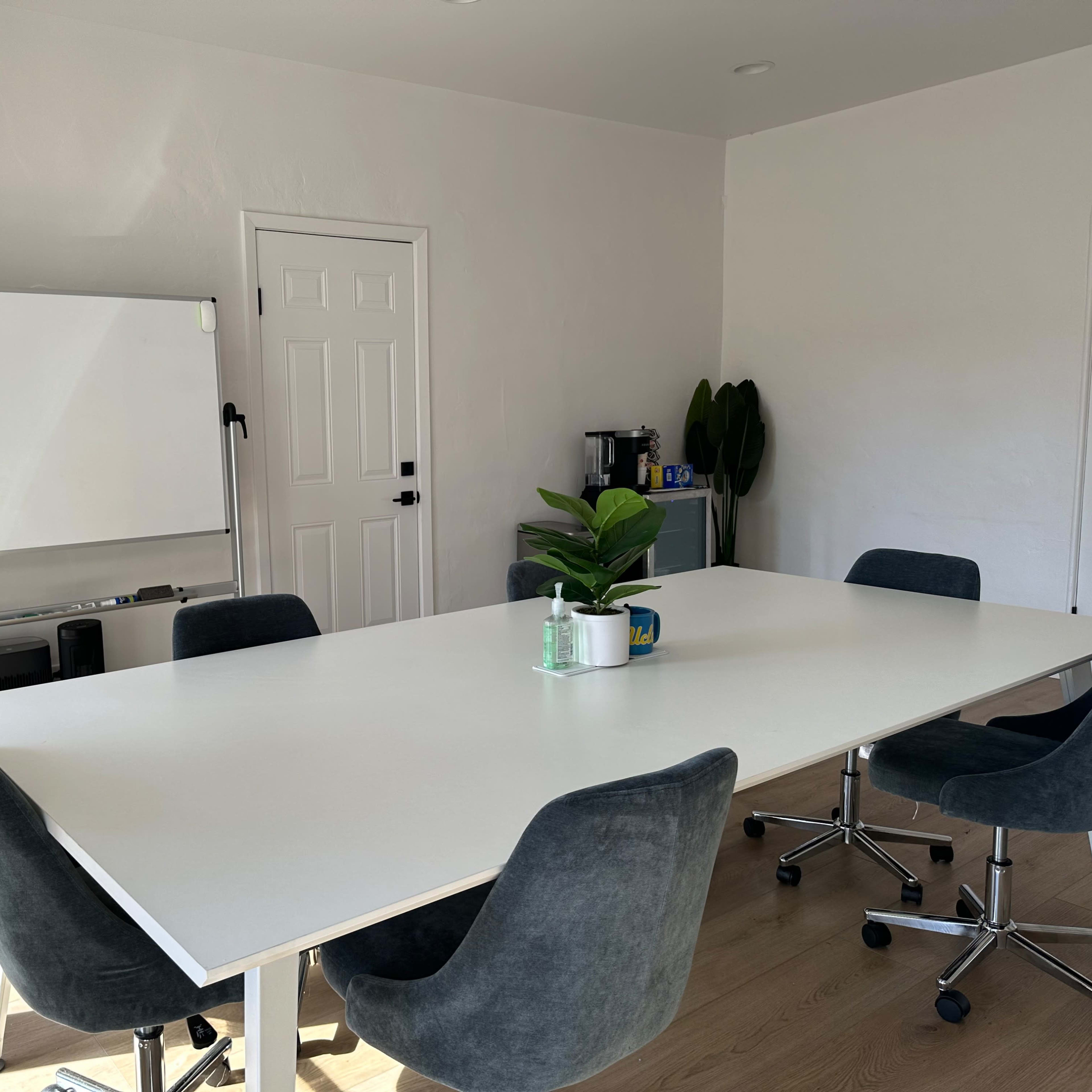 A modern conference room features a large white table surrounded by gray chairs, with a whiteboard and coffee station in the background.