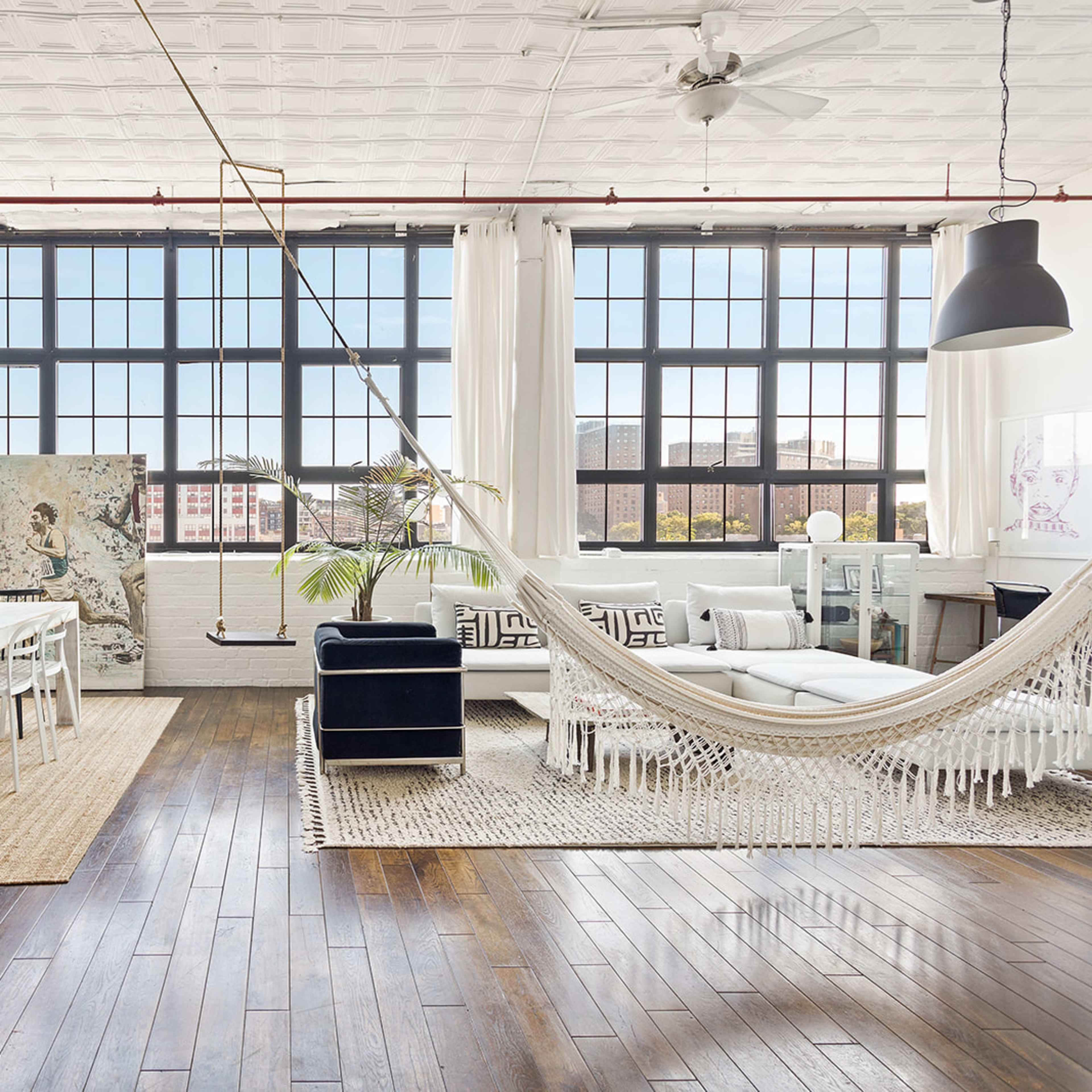 A spacious, well-lit living area features large windows, a dining table, a hammock, and a mix of seating options on hardwood floors.