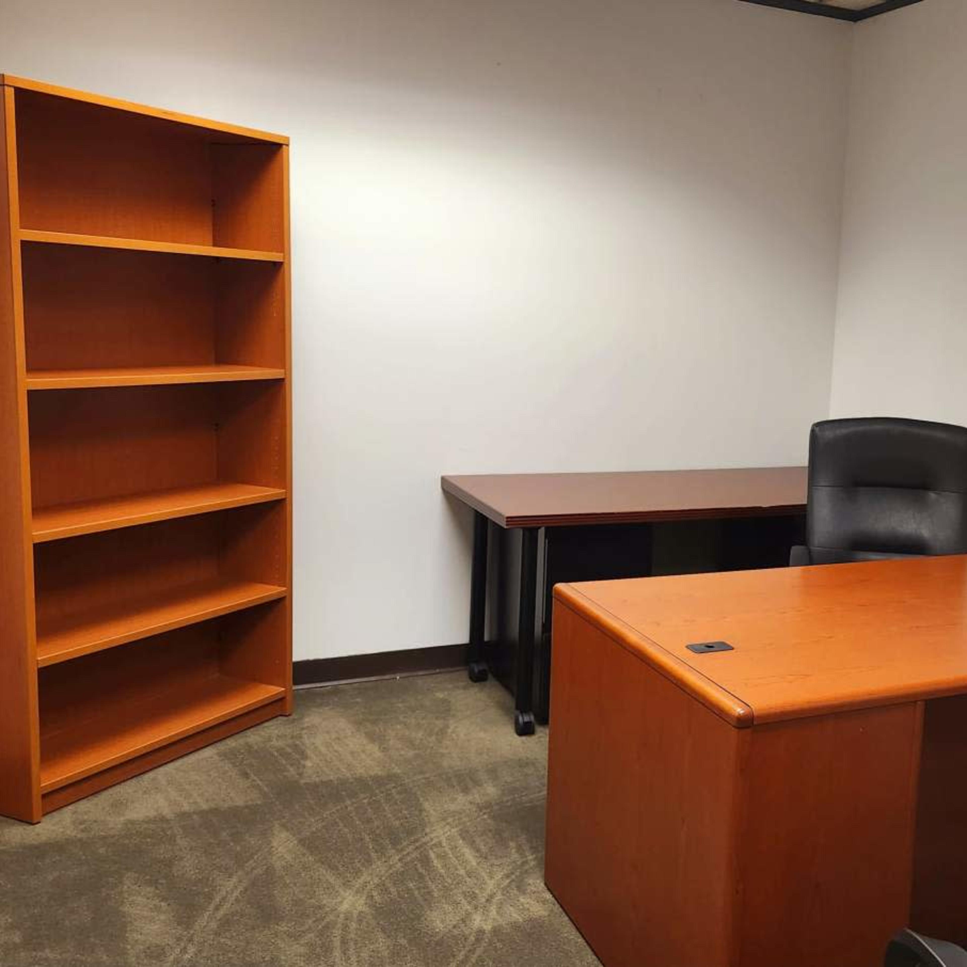An empty office with a wooden desk, a black chair, and a tall bookshelf against the wall.