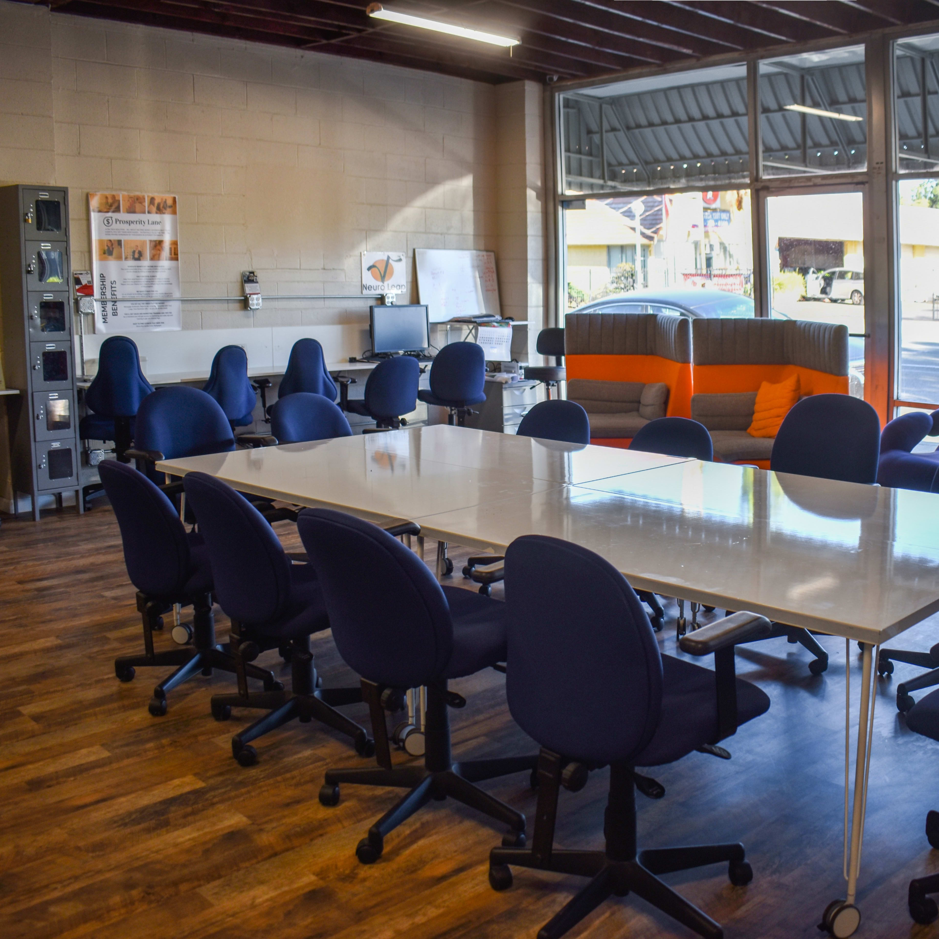 A spacious modern meeting room features a large white table surrounded by blue chairs, with additional seating and workstations visible in the background.