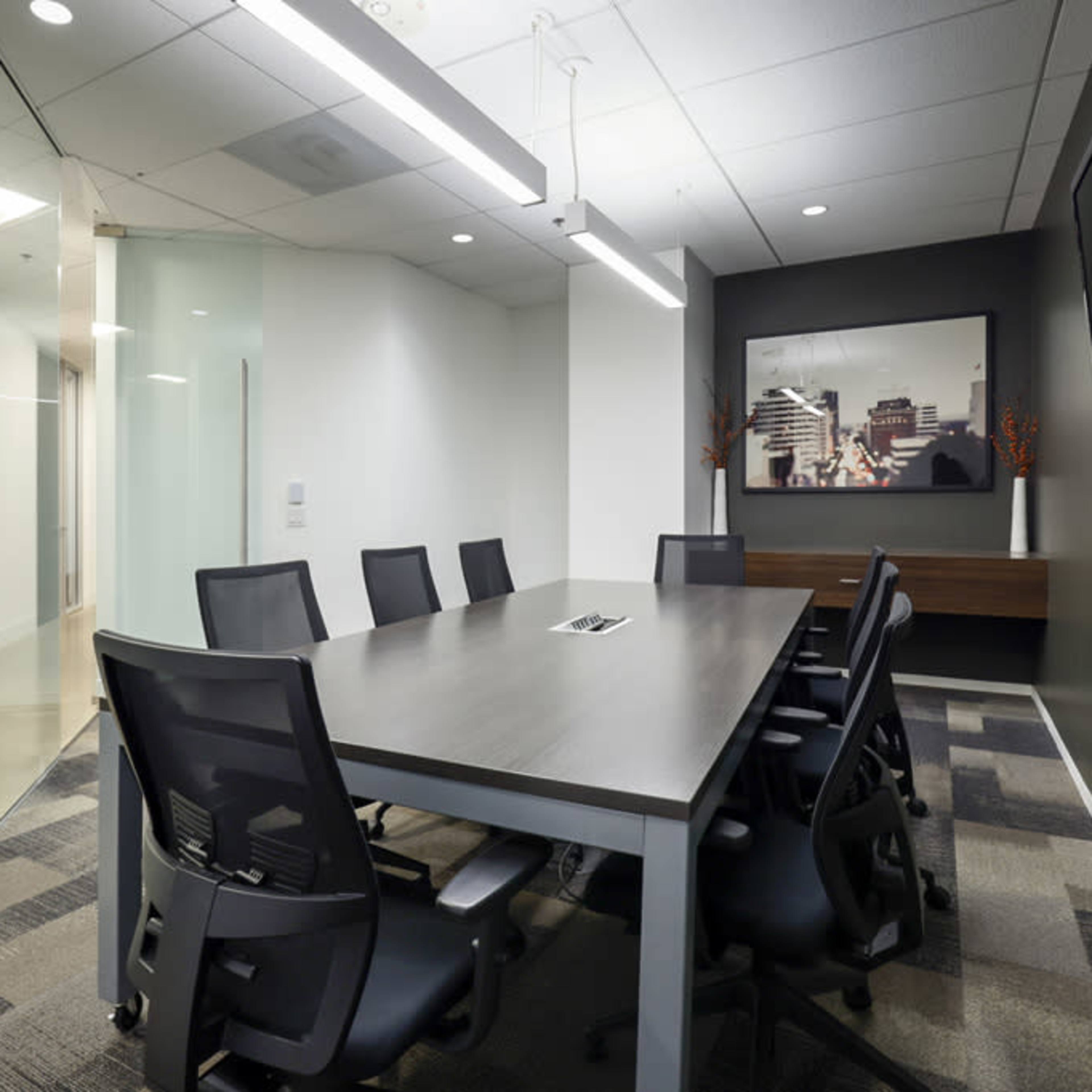 A modern conference room featuring a large table with eight black chairs, a wall-mounted television, and glass walls that lead to an adjacent room.