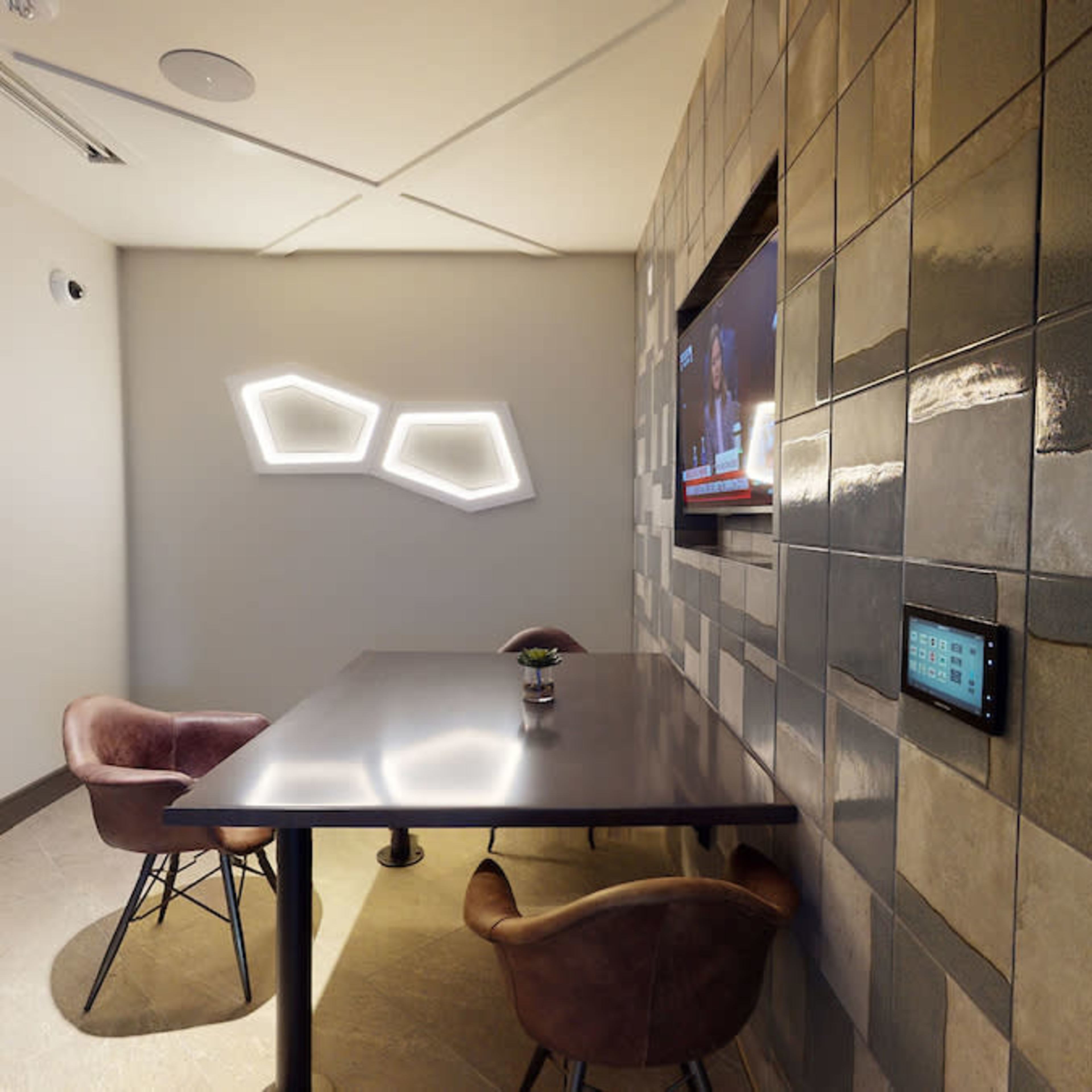 A small meeting room with a dark table, two chairs, a wall featuring geometric decorative lights, and a television mounted on the wall.