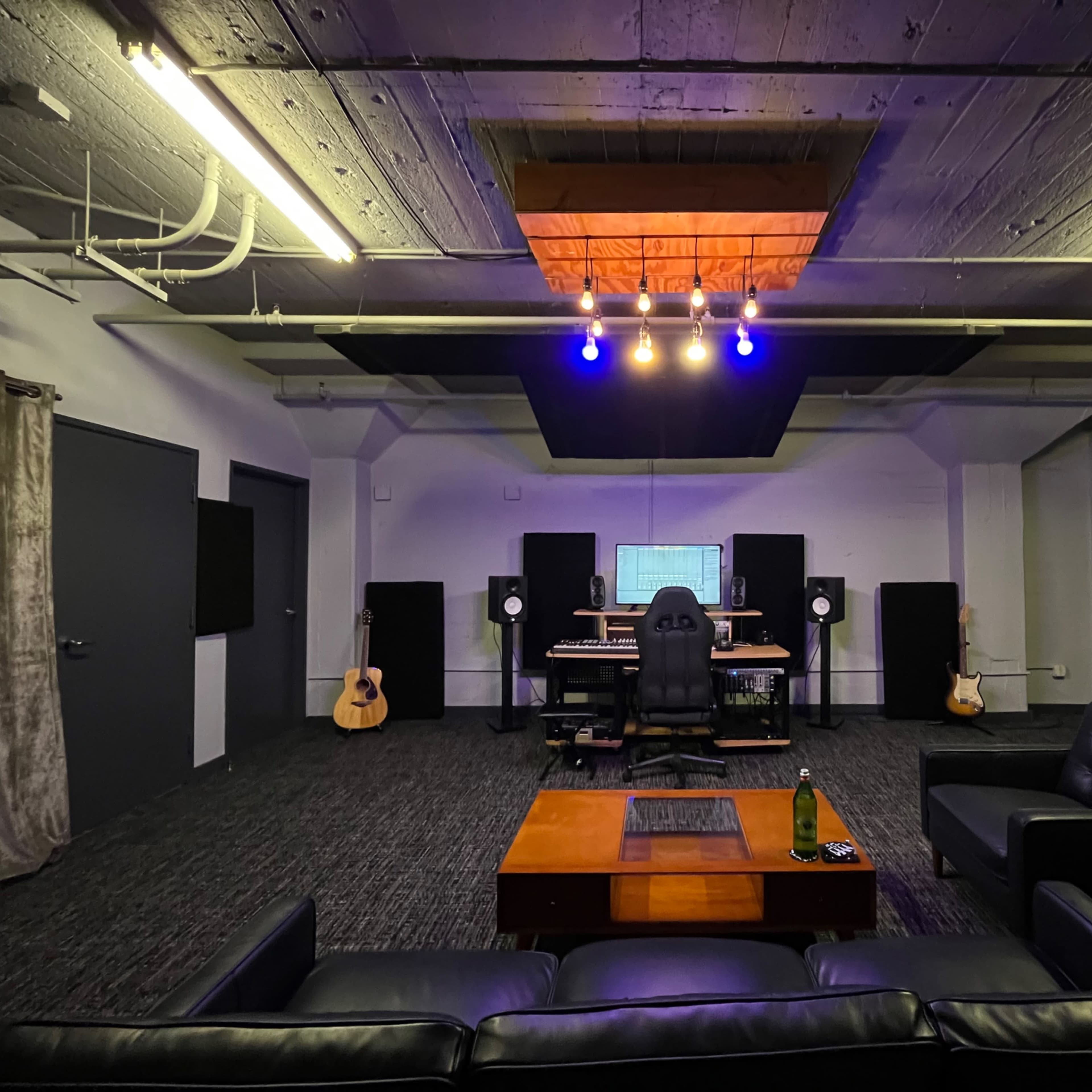 A music studio featuring a central control desk with audio equipment, surrounded by acoustic panels, two guitars, and a comfortable seating area.
