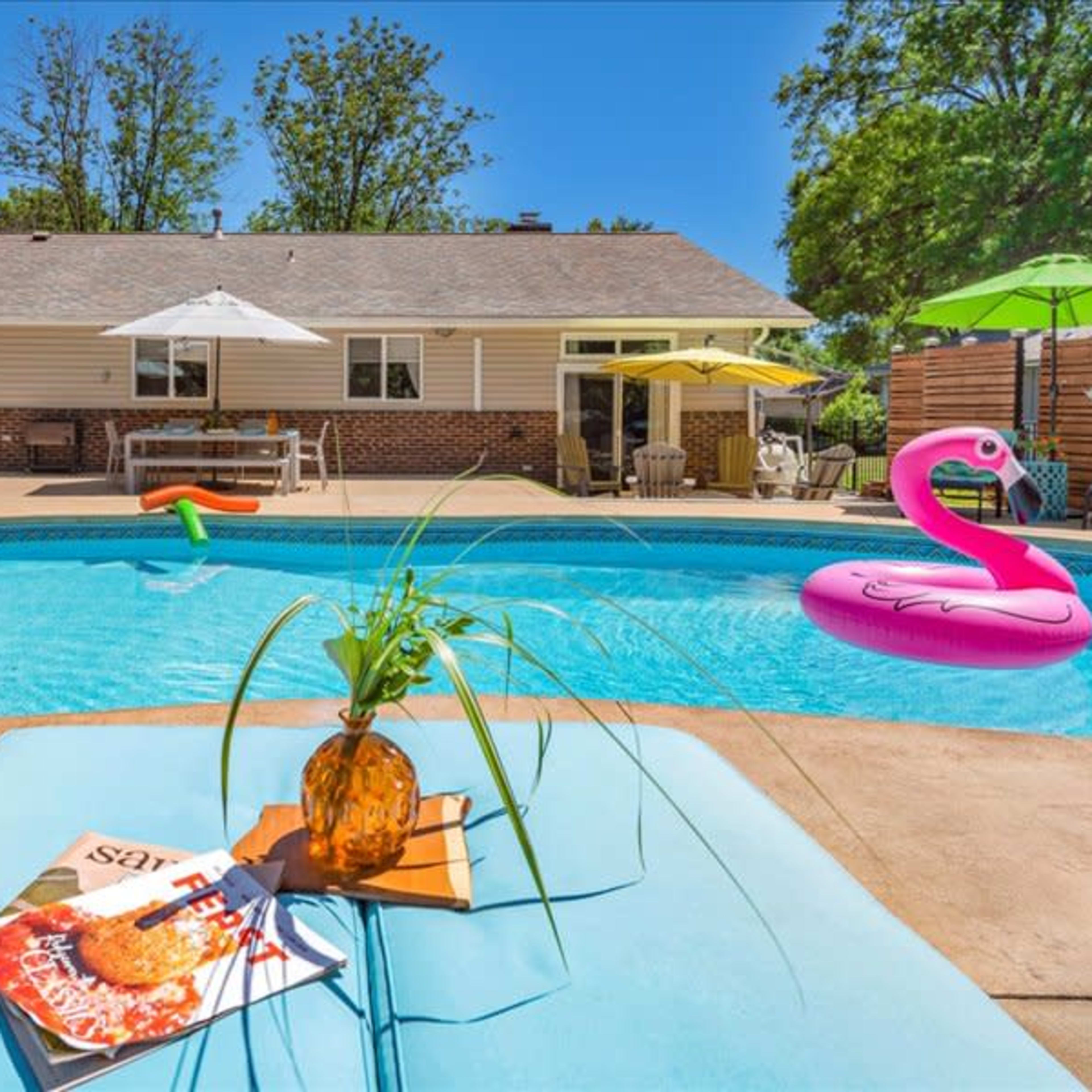 A backyard swimming pool with lounge chairs, a table with a magazine, and a pink inflatable flamingo floating on the water.