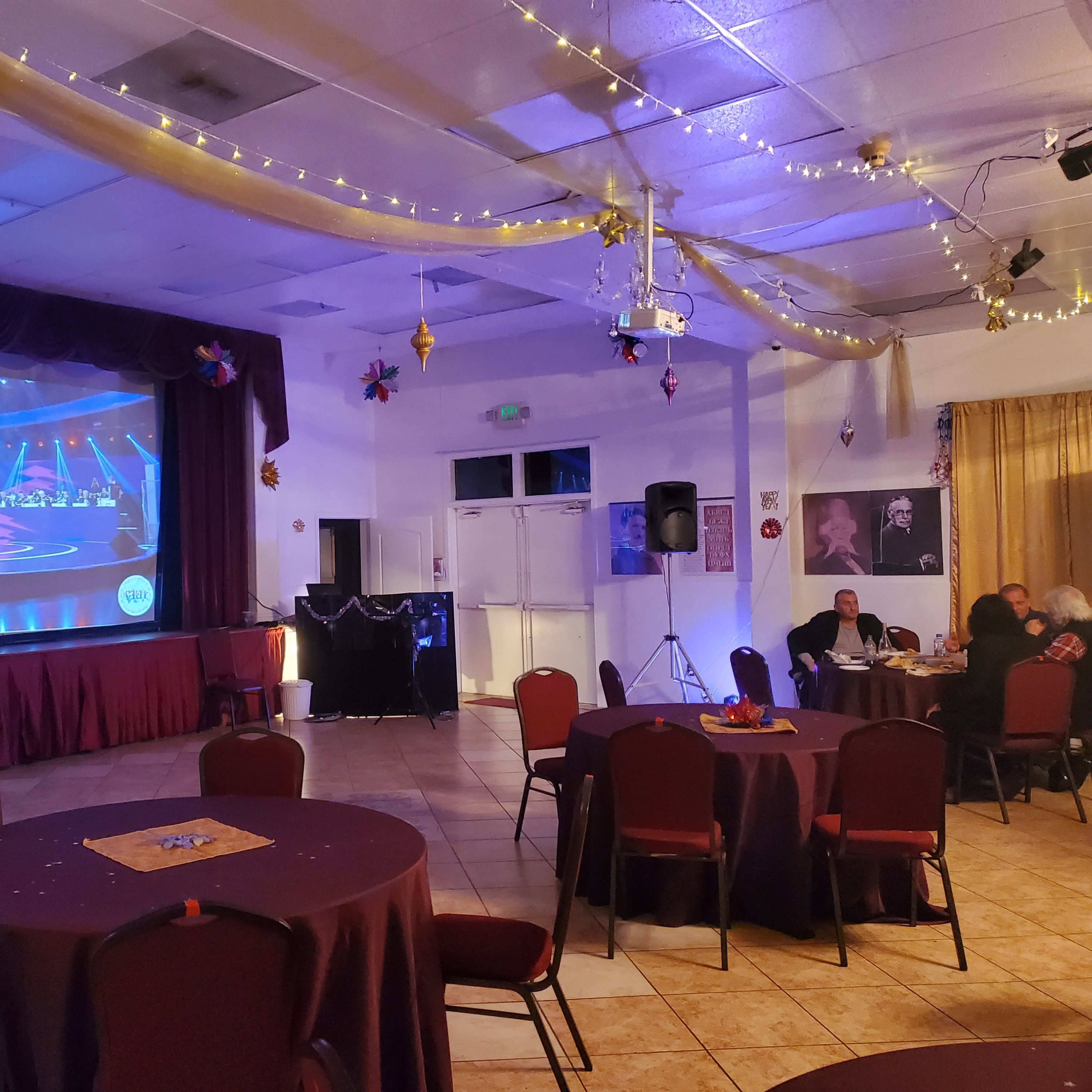 A performer stands on stage in front of a large screen displaying a musical performance, with round tables and chairs arranged in the foreground of the room.