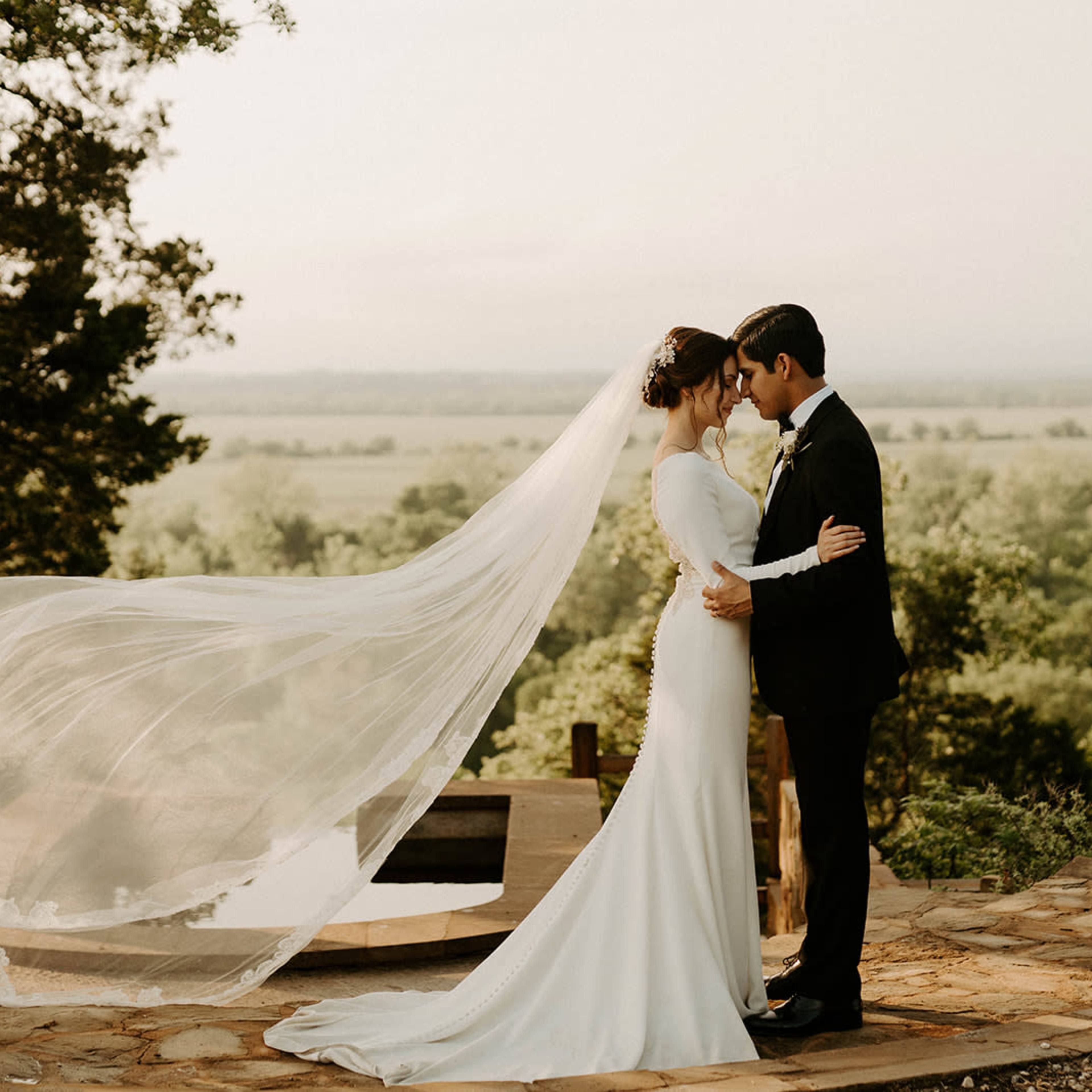 A couple stands embrace on a stone path with a flowing veil billowing in the breeze against a backdrop of lush greenery and a distant horizon.