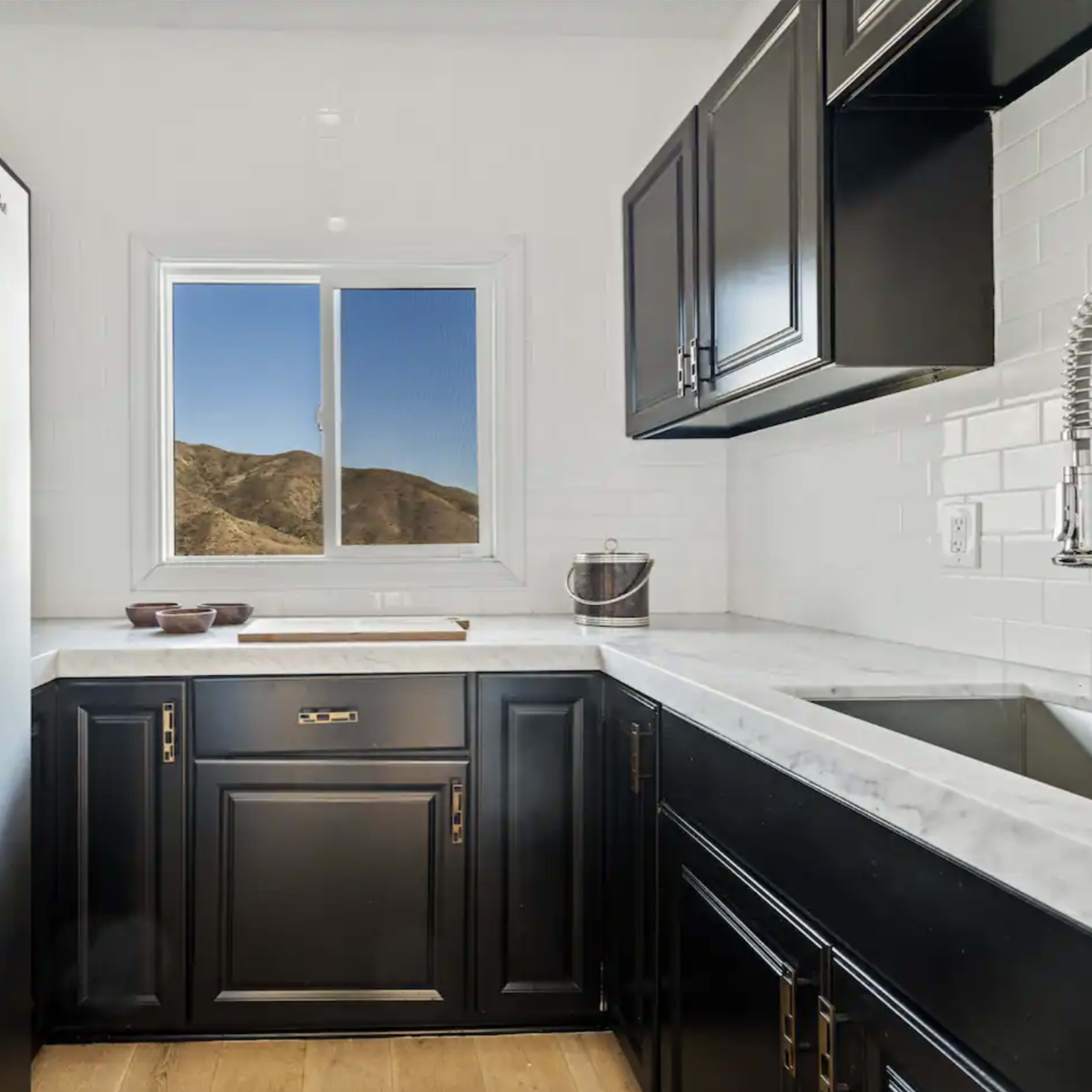 A compact kitchen features black cabinetry, a stainless steel refrigerator, and a window overlooking a mountainous landscape.