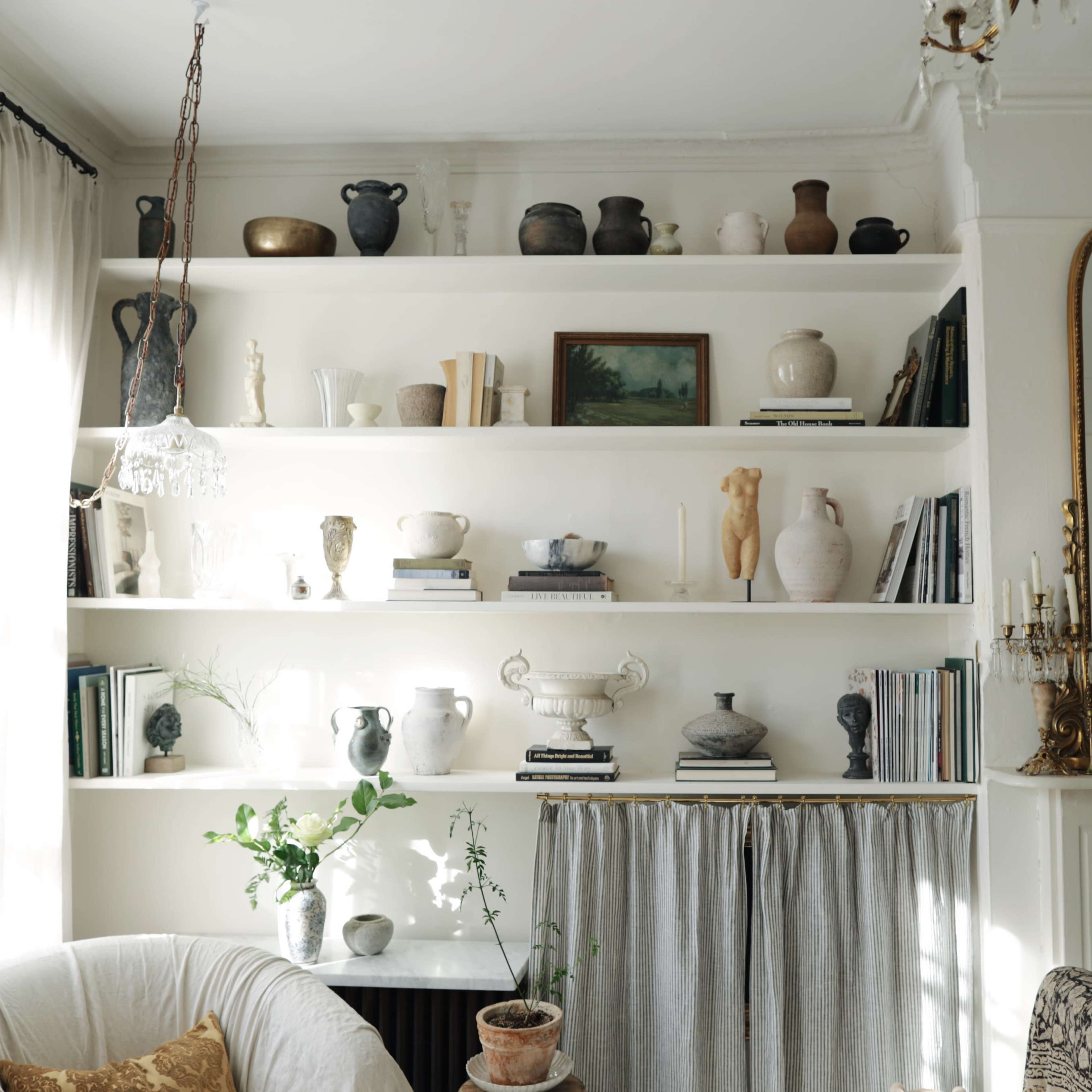 A well-organized shelf displays a variety of ceramic vases, books, and decorative items against a white wall in a softly lit room.