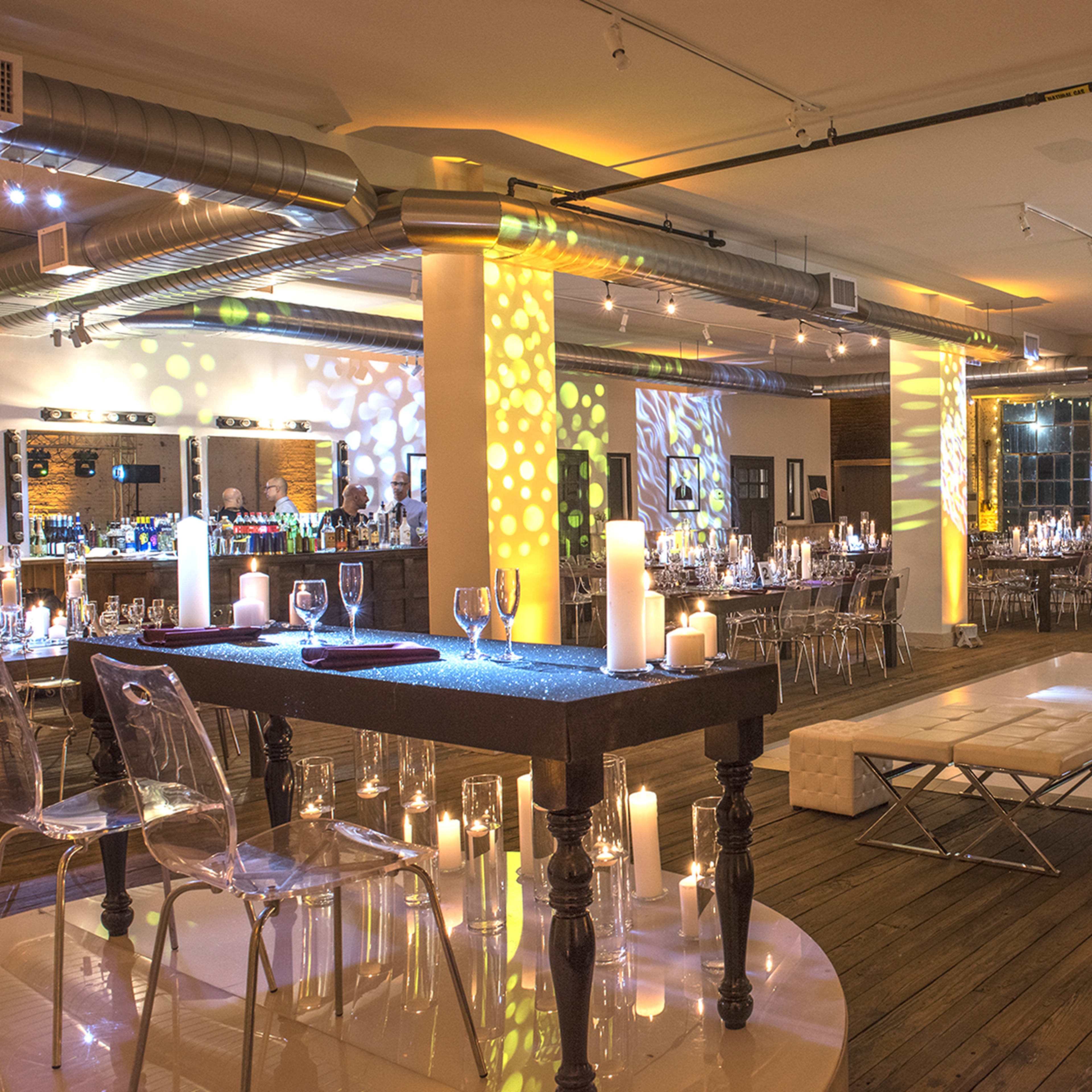 A spacious event venue with elegantly arranged tables, a dance floor in the center, and ambient lighting featuring projections on the walls.