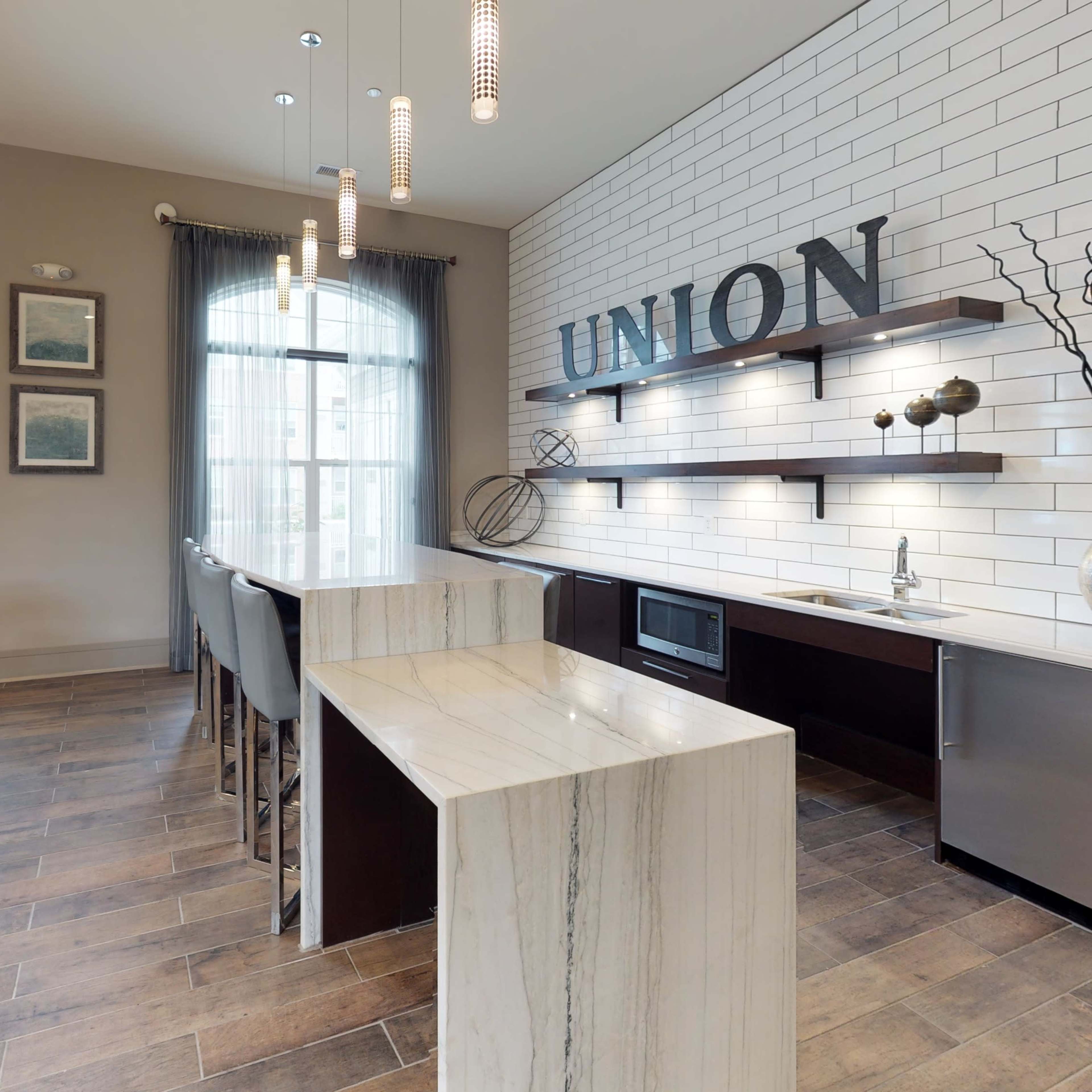 A modern kitchen area features a white tiled wall with "UNION" displayed, a countertop with a sink and microwave, and seating at a high table alongside large windows.