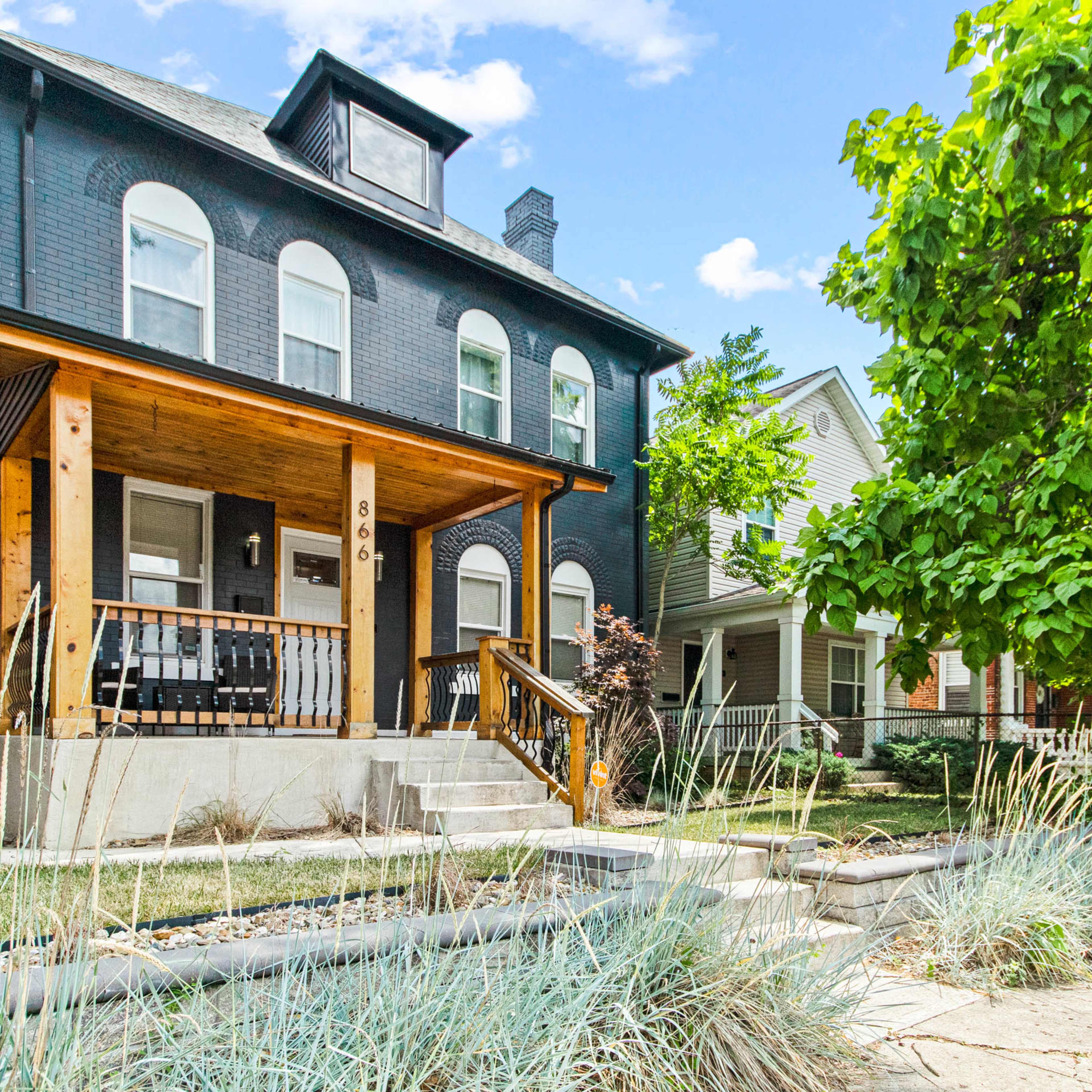 A dark-painted, three-story house with a wooden front porch and steps, surrounded by green foliage and other residential buildings.