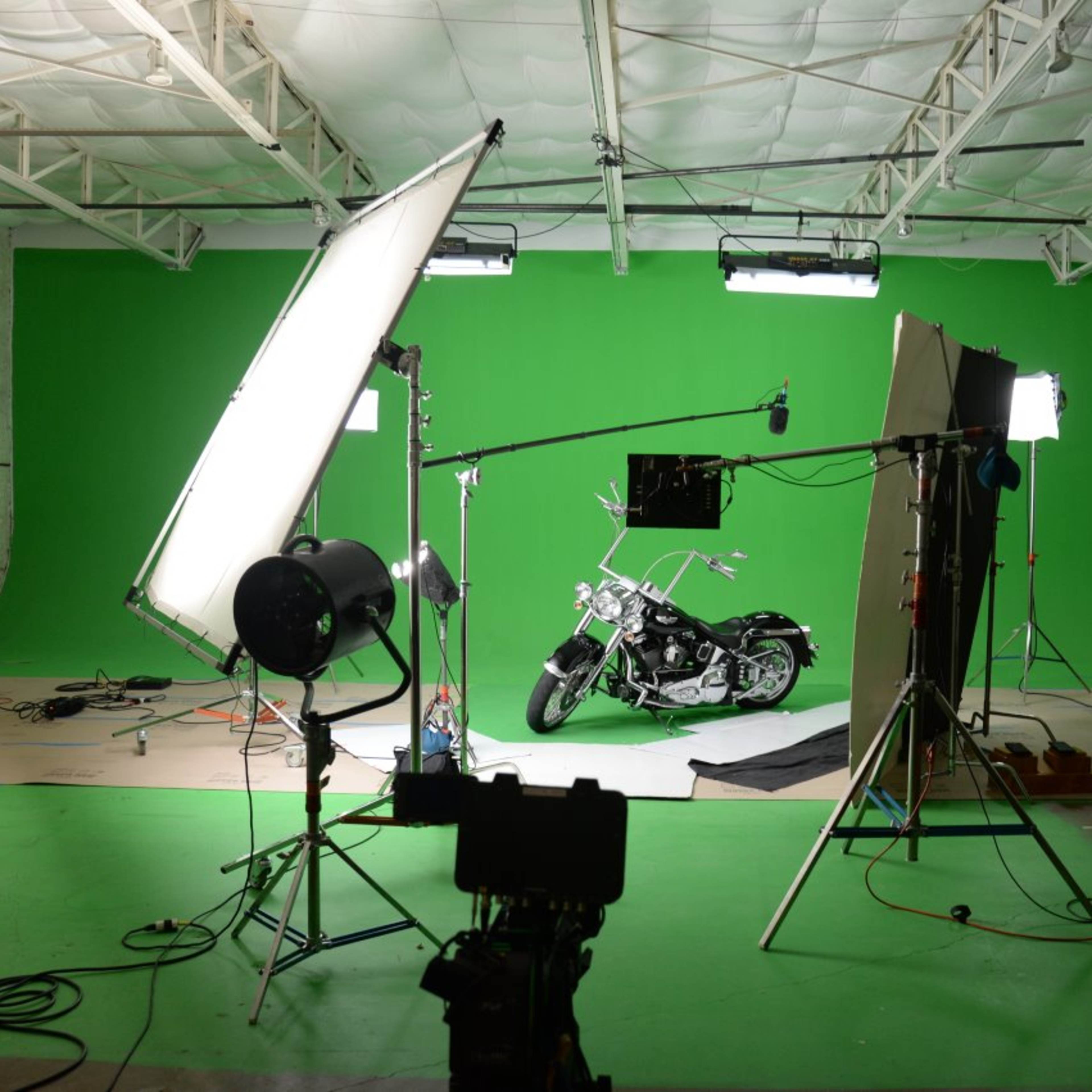 A motorcycle positioned on green flooring in a studio with green backdrop, surrounded by professional lighting and production equipment.