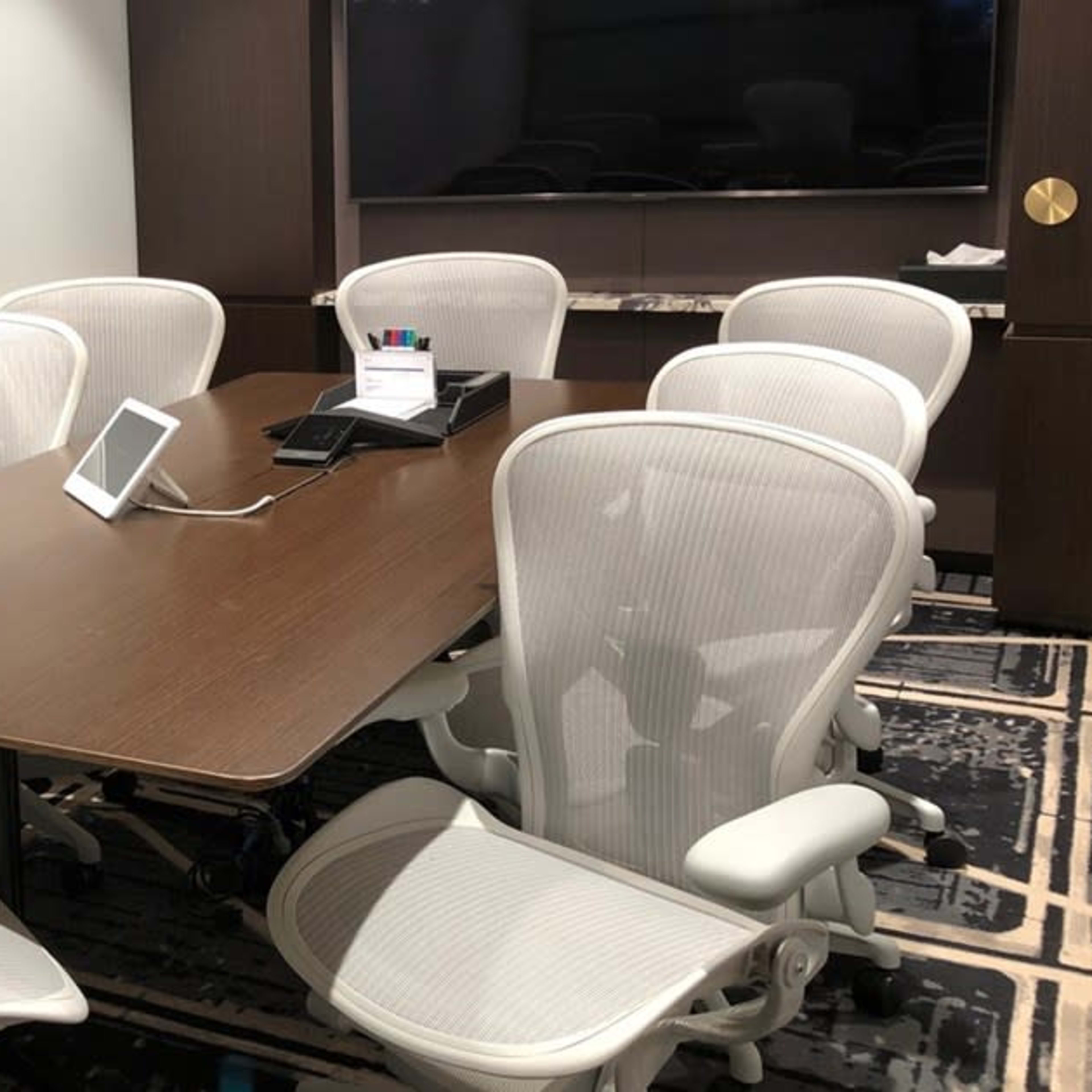 A conference room features a wooden table surrounded by several white office chairs and a screen on the wall.
