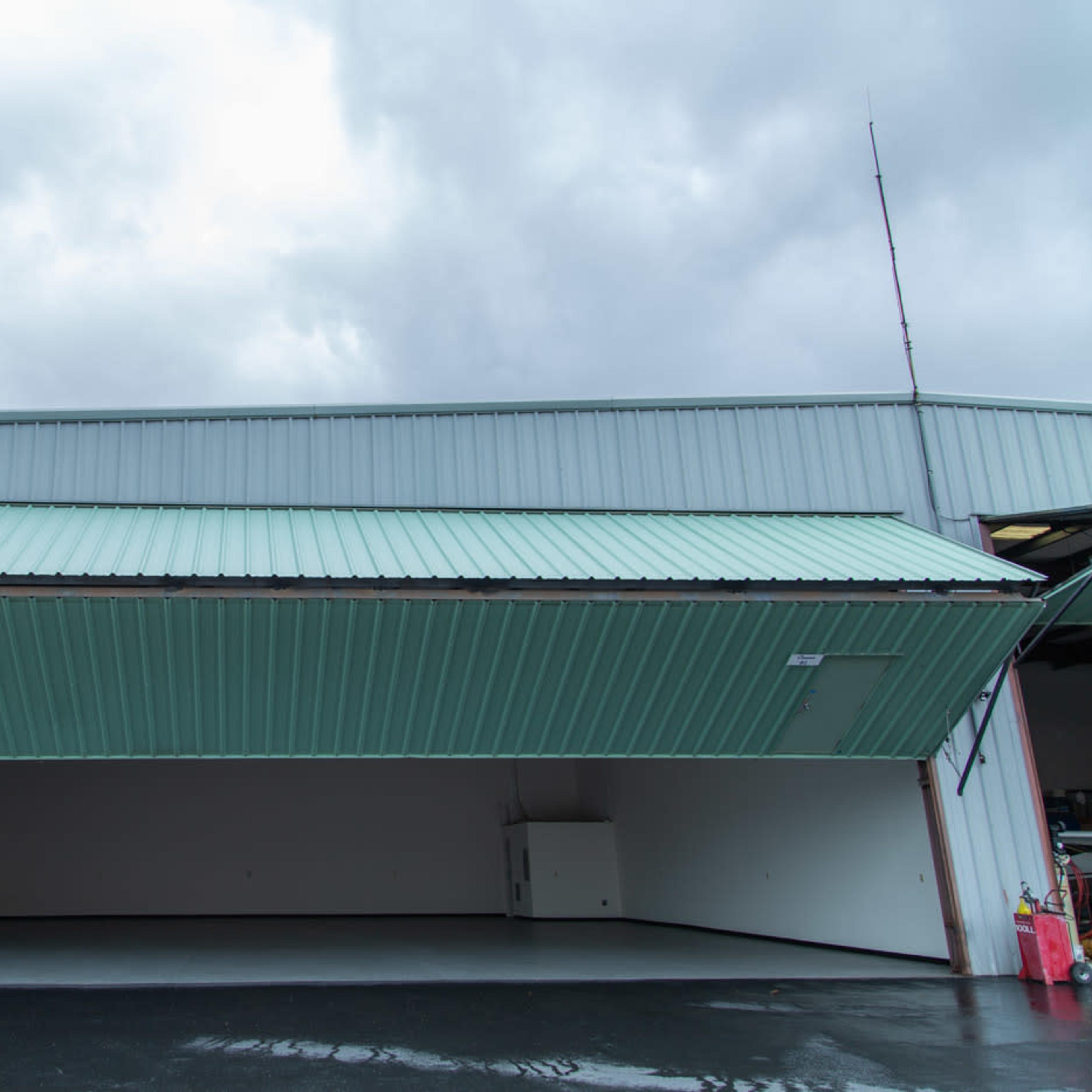 A large hangar with an open door and a green corrugated roof is shown against a cloudy sky.