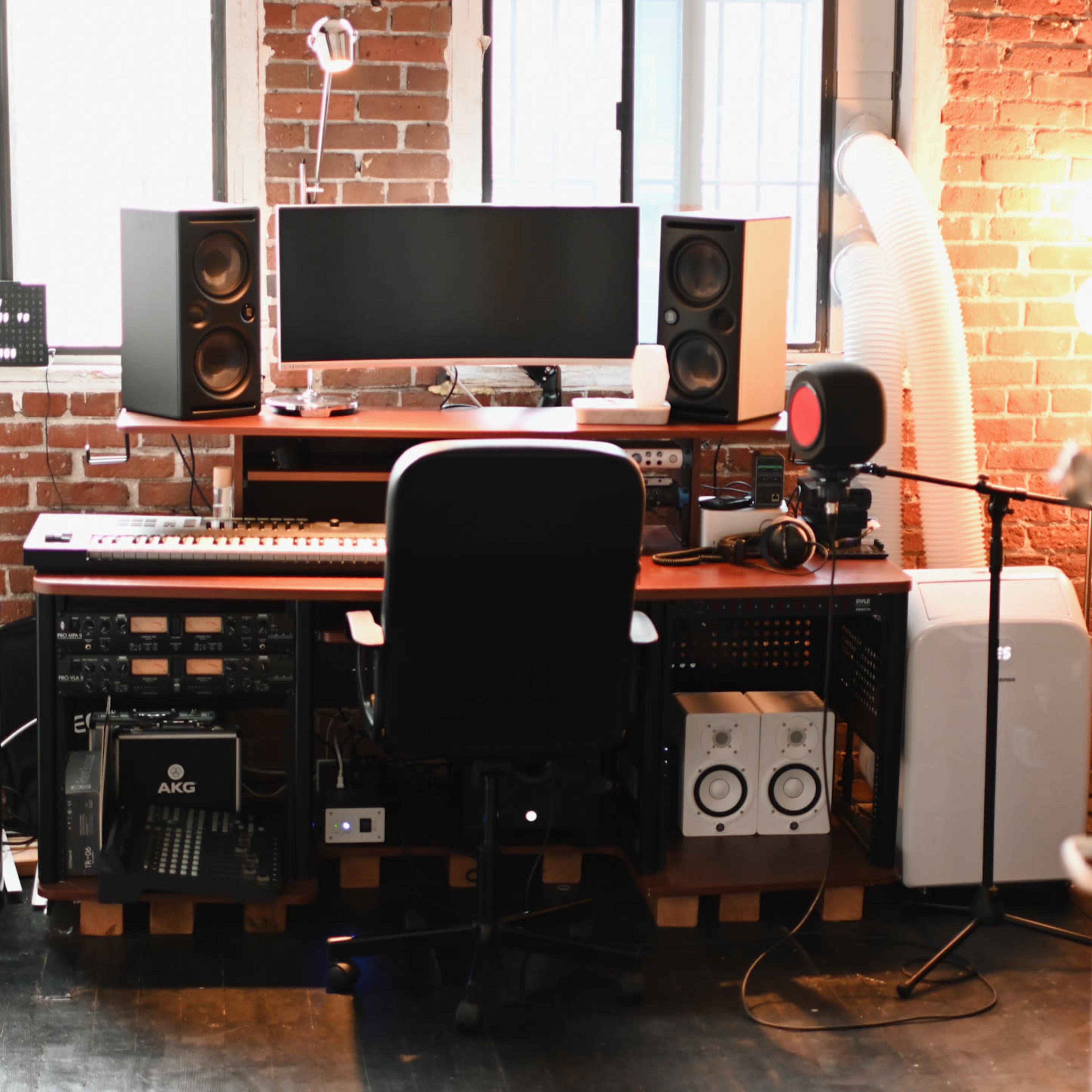 A music production studio setup featuring a desk with a computer monitor, speakers, a keyboard, and various audio equipment against a brick wall.