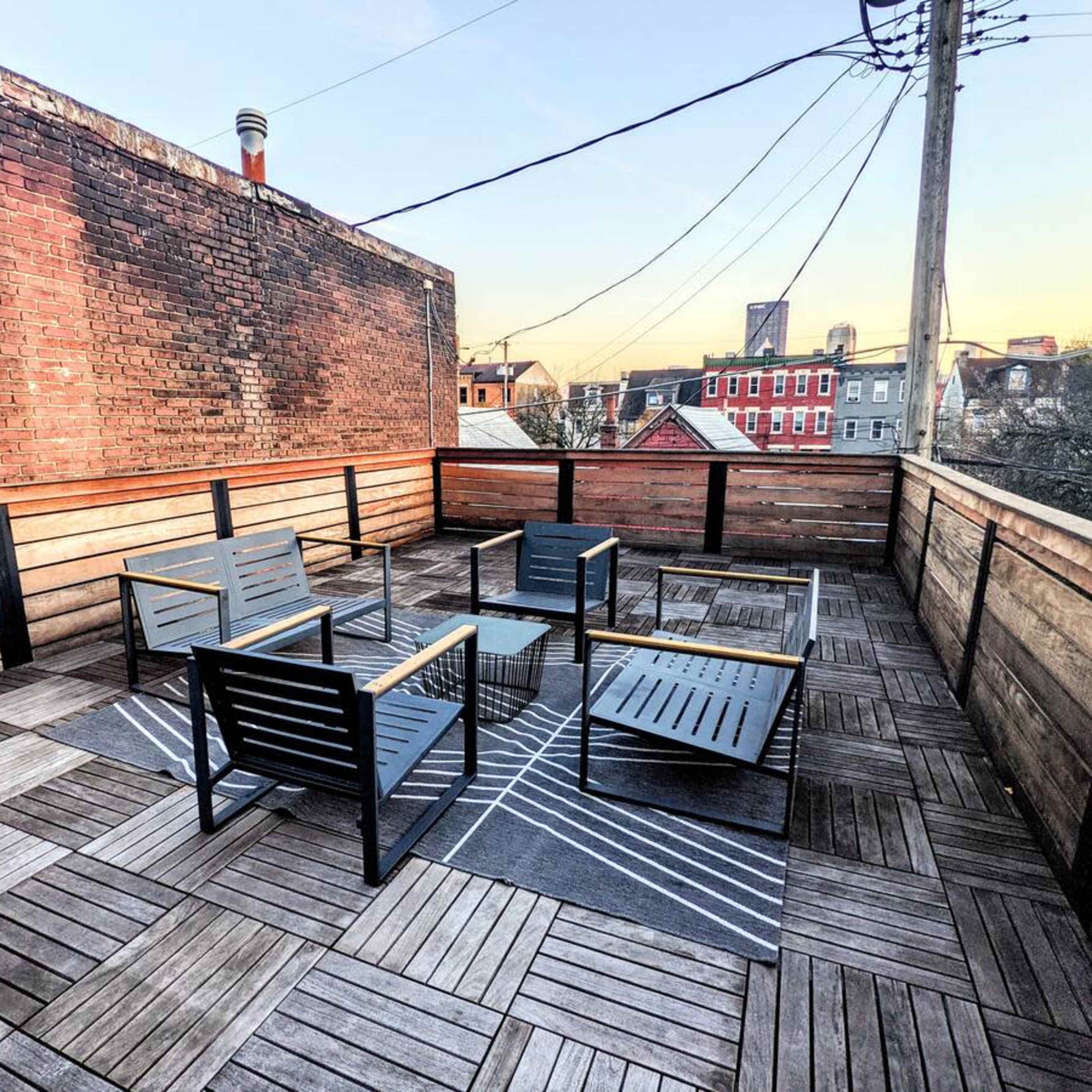 A rooftop terrace with wooden decking and metal-framed seating arranged around a central table, set against a backdrop of urban buildings and power lines.