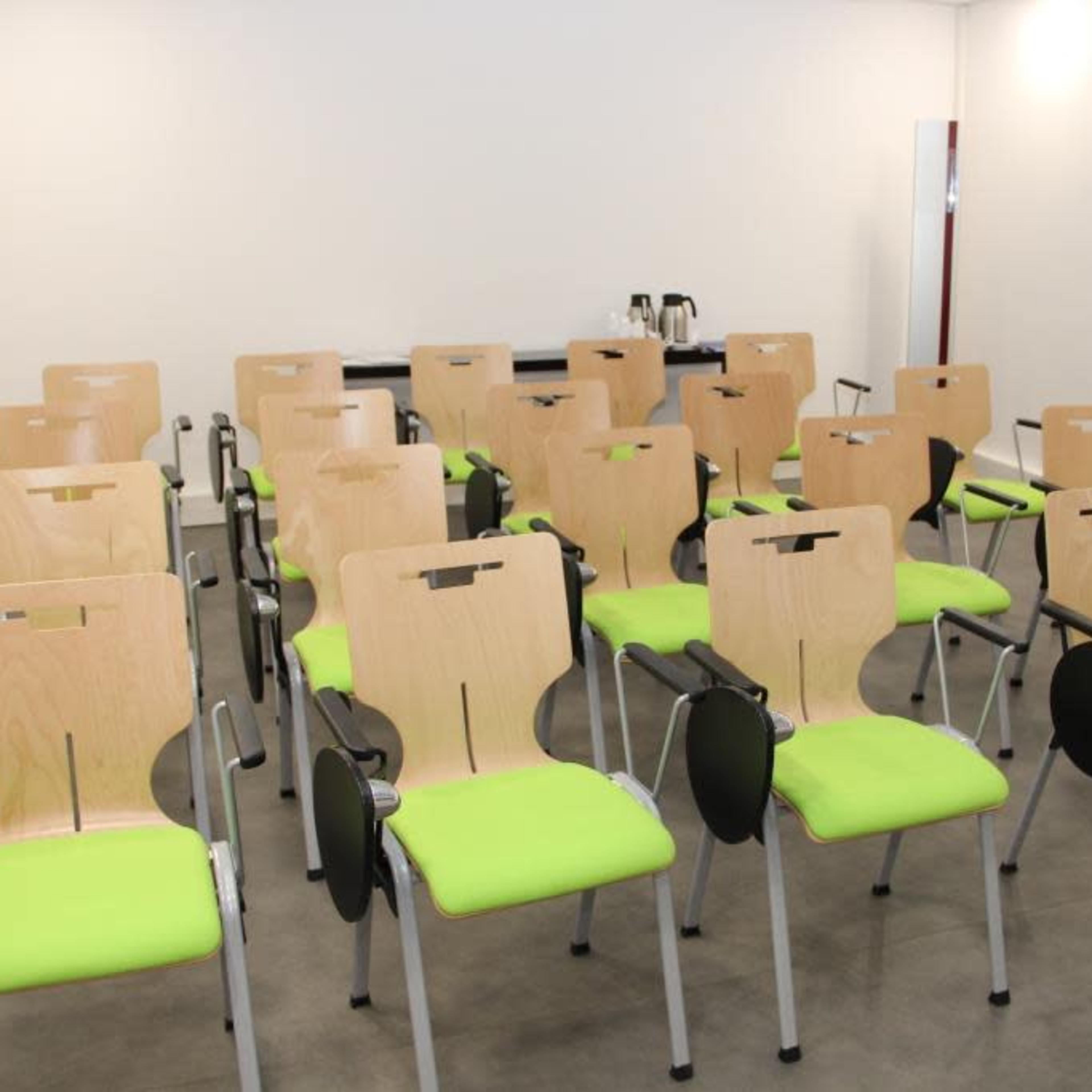 A row of empty chairs with wooden backs and green cushions is arranged neatly in a bright room.