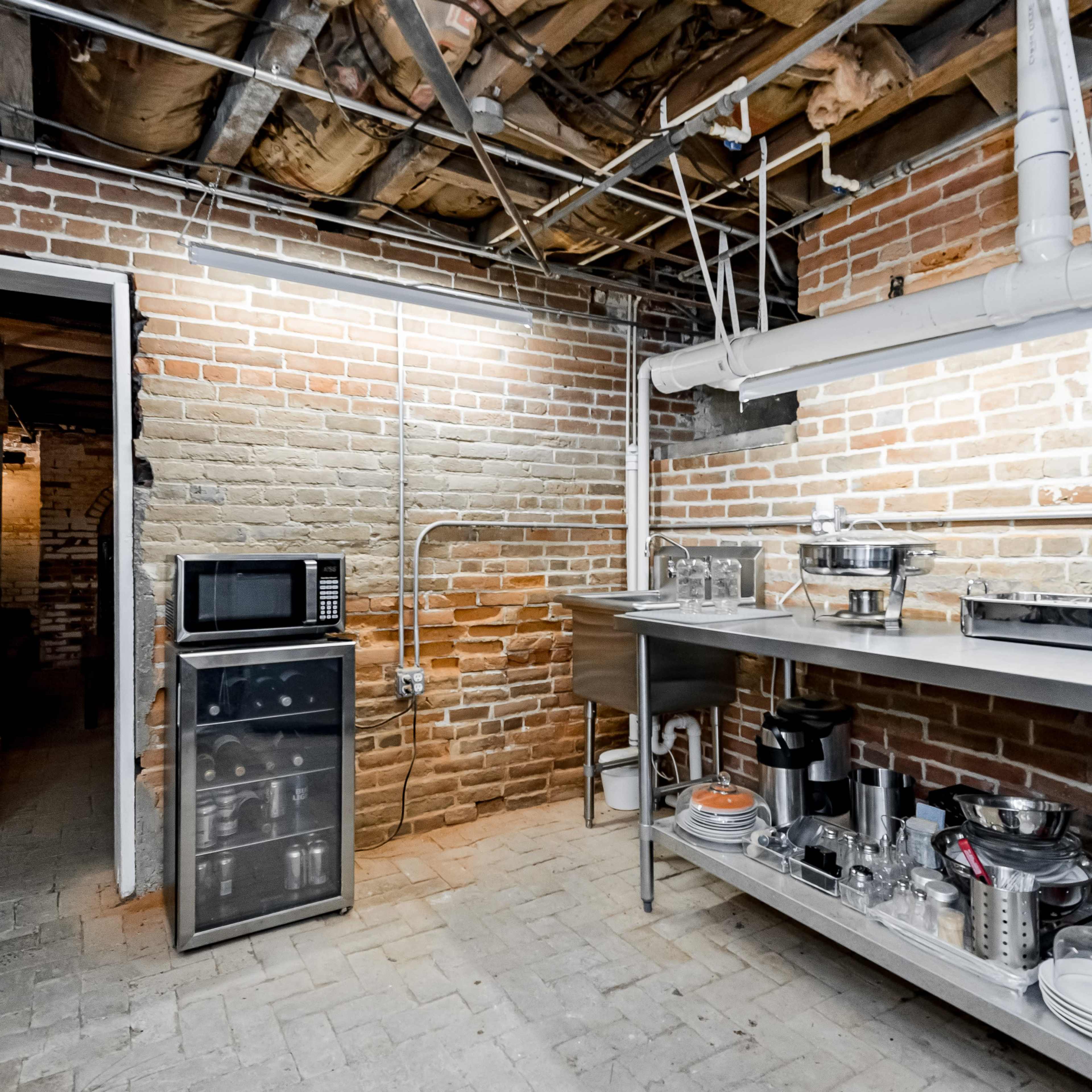 A basement kitchen area with exposed brick walls, a microwave, a mini-fridge, and various kitchen utensils on a stainless steel countertop.