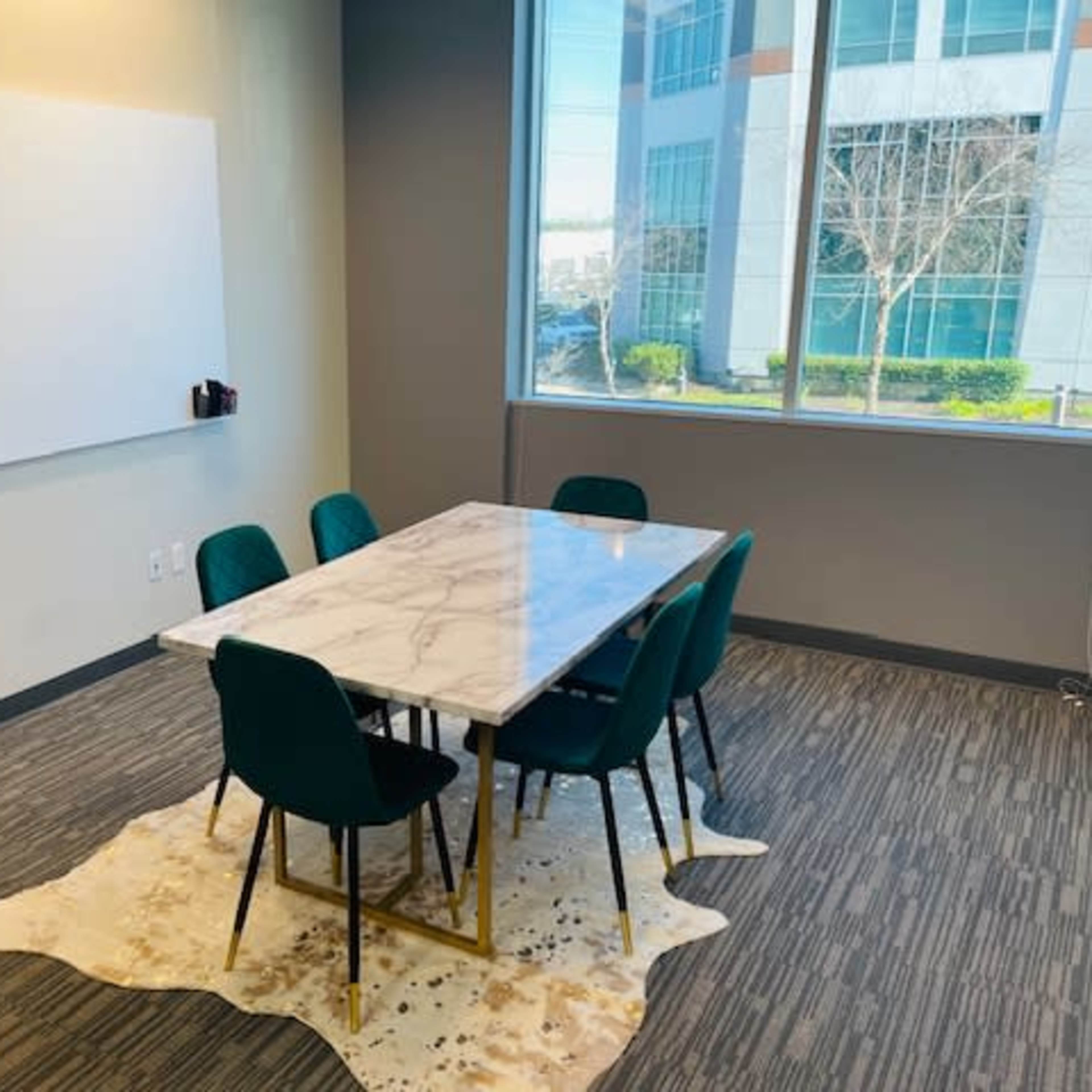 A modern meeting room features a marble-top table surrounded by green chairs, with a large whiteboard on one wall and a window overlooking an office building.